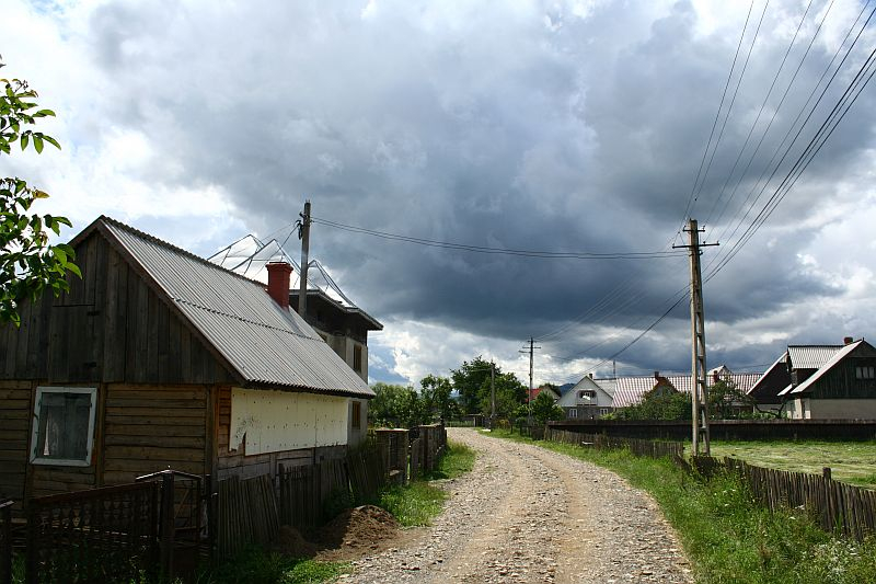 A rustic dirt road leads through a tranquil village in Romania with wooden houses under a dramatic sky, hinting at an approaching storm.