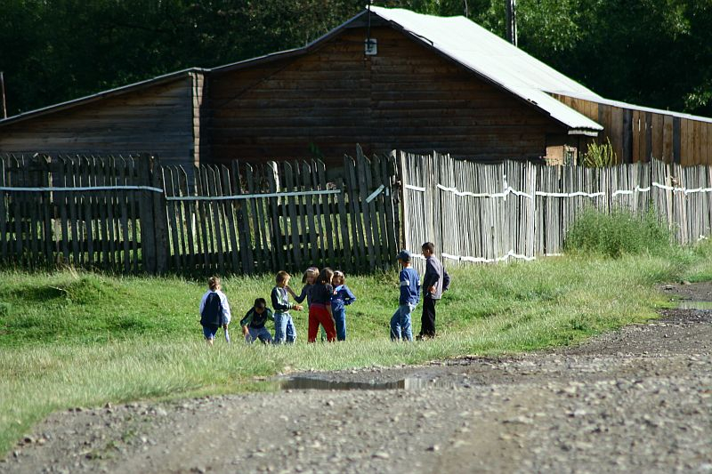 A group of children walk beside a wooden fence in a rural Romanian setting, with wooden houses in the background and a gravel road in the foreground, suggesting a communal or village lifestyle.