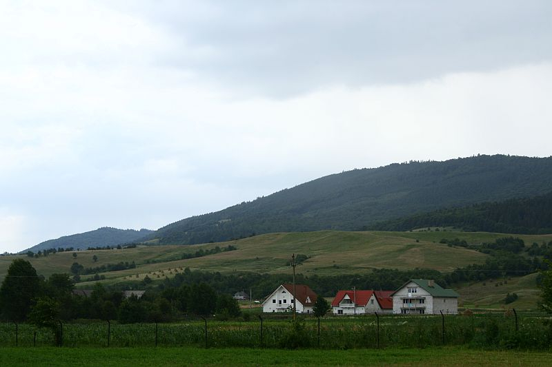 A serene Romanian countryside scene with rolling hills in the background, a cluster of houses in the foreground, and an overcast sky hinting at the possibility of rain.