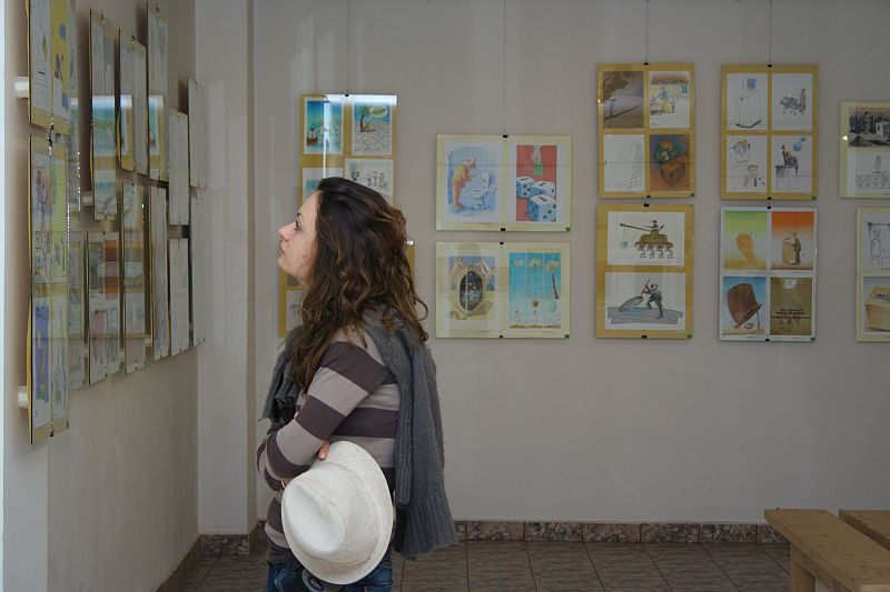 A woman with curly hair, holding a white hat, contemplatively observes colorful artwork displayed in simple frames on a gallery wall in Romania, illuminated by soft lighting.