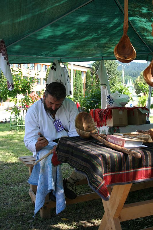 A craftsman in traditional Romanian attire meticulously carves wood under a canopy at an outdoor market, surrounded by artisanal creations, symbolizing cultural heritage and the enduring artistry of handcrafting.