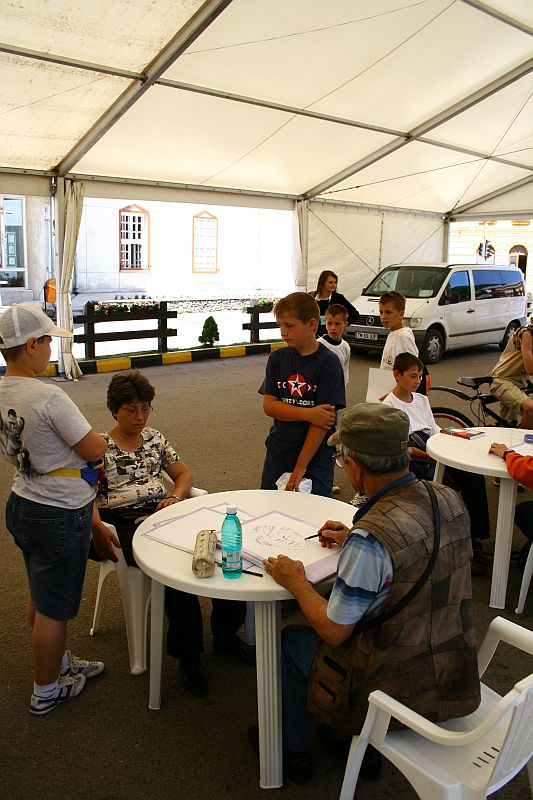 Children gather around a table at an outdoor workshop in Romania, engaging attentively with an instructor who is demonstrating or instructing, encouraging interactive learning in a casual, tented setting.