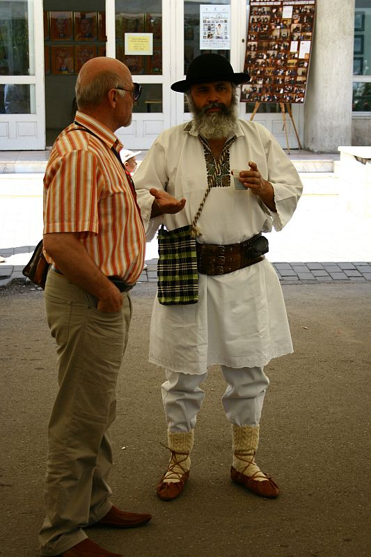 Two men are standing and talking. The man on the left wears glasses, a short-sleeved orange-striped shirt, beige pants, and brown shoes. The man on the right, with a white beard, is dressed in traditional Romanian folk attire: a white shirt, pants, a dark hat, and traditional footwear.