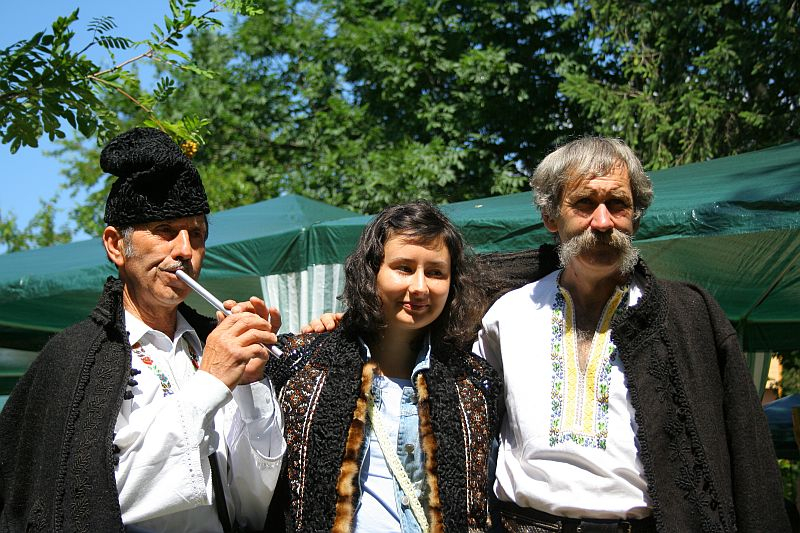 Three individuals in traditional Romanian attire—two men with vests and furry hats flank a smiling woman in a blouse—pose outdoors on a sunny day, with the elder man on the right holding a wooden flute