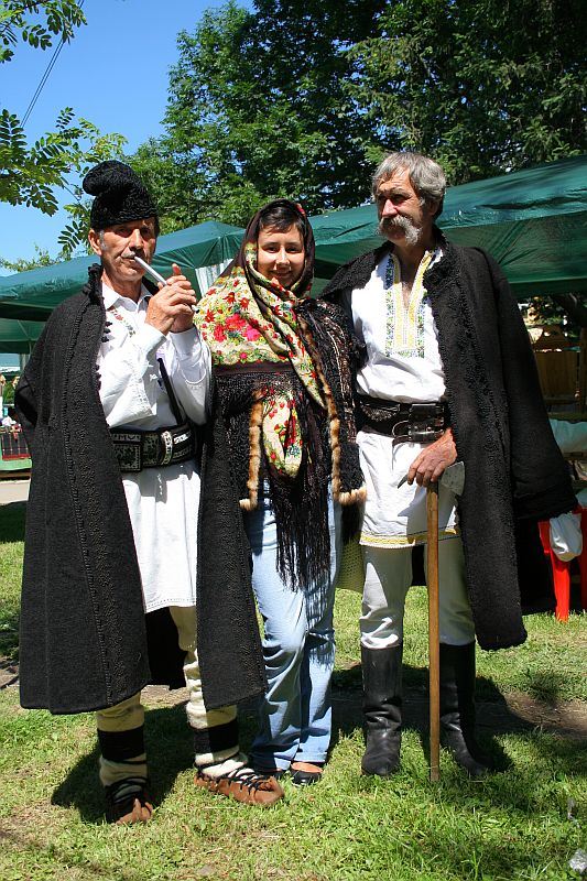 Three individuals in traditional Romanian attire pose outdoors; two men in embroidered vests and fur hats flank a woman in a colorful headscarf, exemplifying cultural heritage.