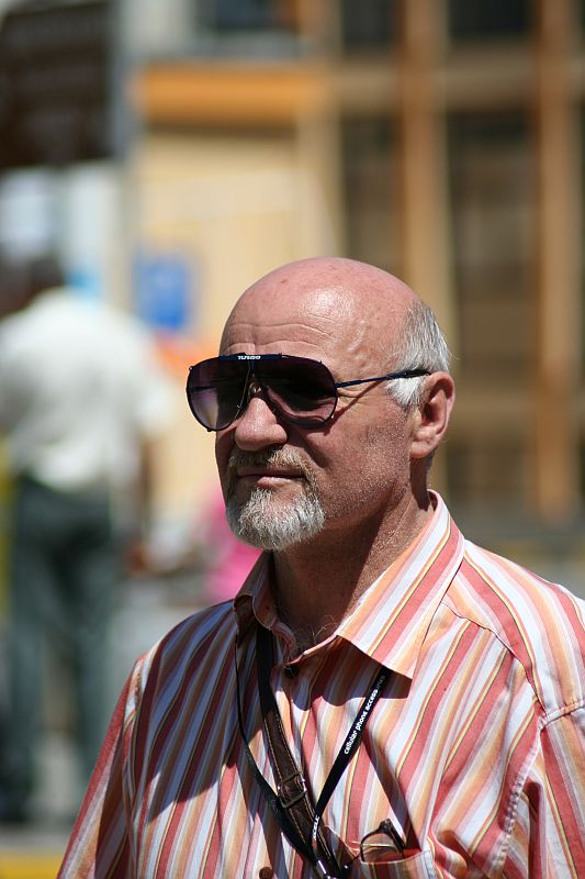A man with a mustache wearing sunglasses and a striped shirt stands in focus, with a blurred background indicating a bustling outdoor setting in Romania.