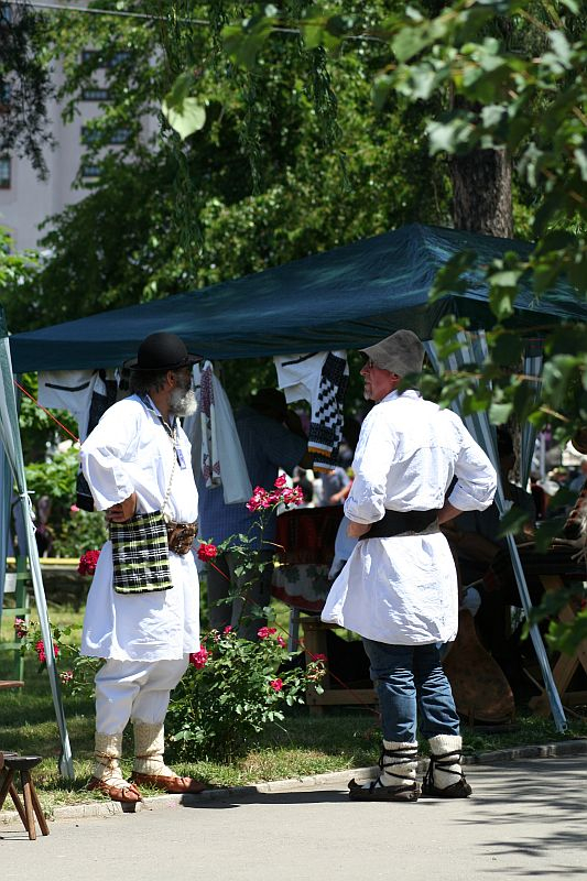 Two individuals in traditional Romanian folk costumes engage in a conversation at a sunny outdoor market, surrounded by tents and handicrafts.