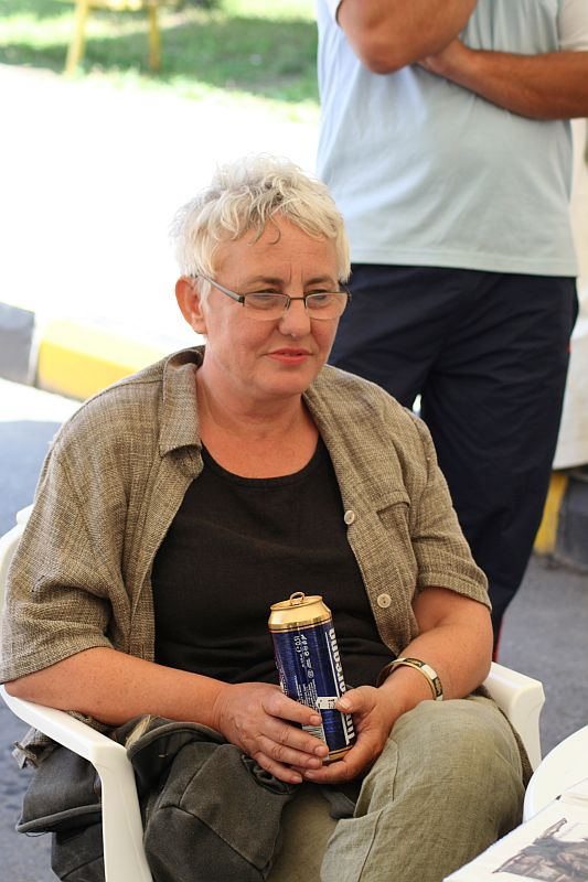 A woman with short, light-colored hair, wearing glasses and a checkered jacket, casually sits holding a can, attentively listening with a slight smile in a tented area in Romania.