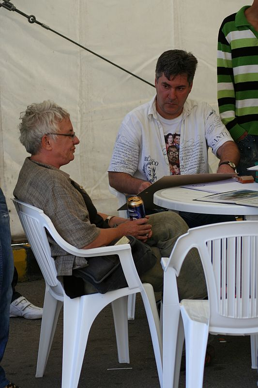 Two individuals engaged in conversation at a casual gathering in Romania, with one sitting across a table holding a beverage can, surrounded by a relaxed atmosphere under a tented structure.