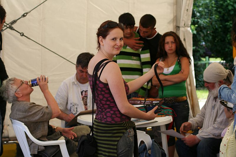 A woman stands smiling at the camera, holding a bag, in a tent in Romania where people are engaged in various activities, some sitting and others conversing, suggesting a casual gathering or event.