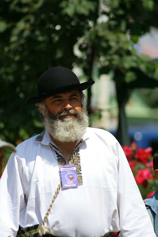 A man with a full beard and a black hat, wearing a traditional Romanian folk shirt adorned with a badge, gazes into the distance at an outdoor event.