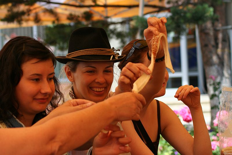 Three people joyfully stretch a piece of cheese, displaying its stringy texture, while sharing a moment of camaraderie around a table with food and flowers in the background in Romania.