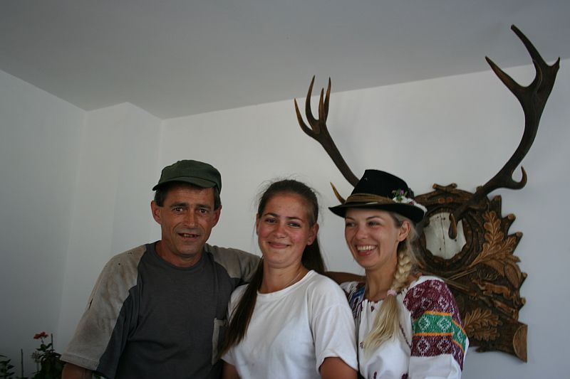 Three individuals pose smiling: a man in a cap, a young woman in white, and another with a traditional Romanian hat, with an antler display and clock in the background.