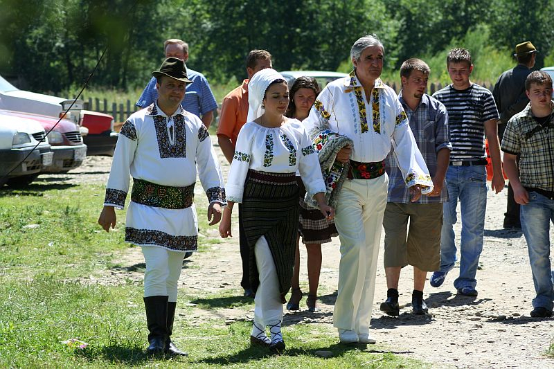 A group of people, some dressed in traditional Romanian embroidered outfits, walk together in the sunshine at a cultural event with nature and parked cars in the background.