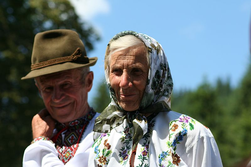 An elderly couple in traditional Romanian attire, with the woman wearing a floral headscarf and embroidered shirt, stands close together against a sunny, tree-lined backdrop, evoking a sense of cultural heritage and
