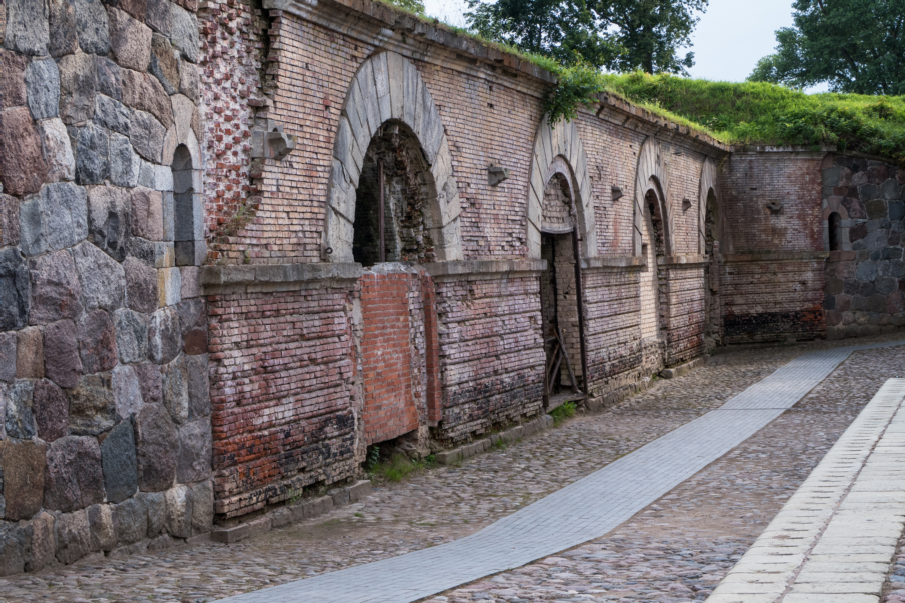 The Daugavpils fortress boasts a historical wall of weathered brick and stone. Arched doorways, partially open, lead into shadowed entries. A cobblestone path, edged by a narrow concrete strip, guides visitors while lush greenery crowns the structure.