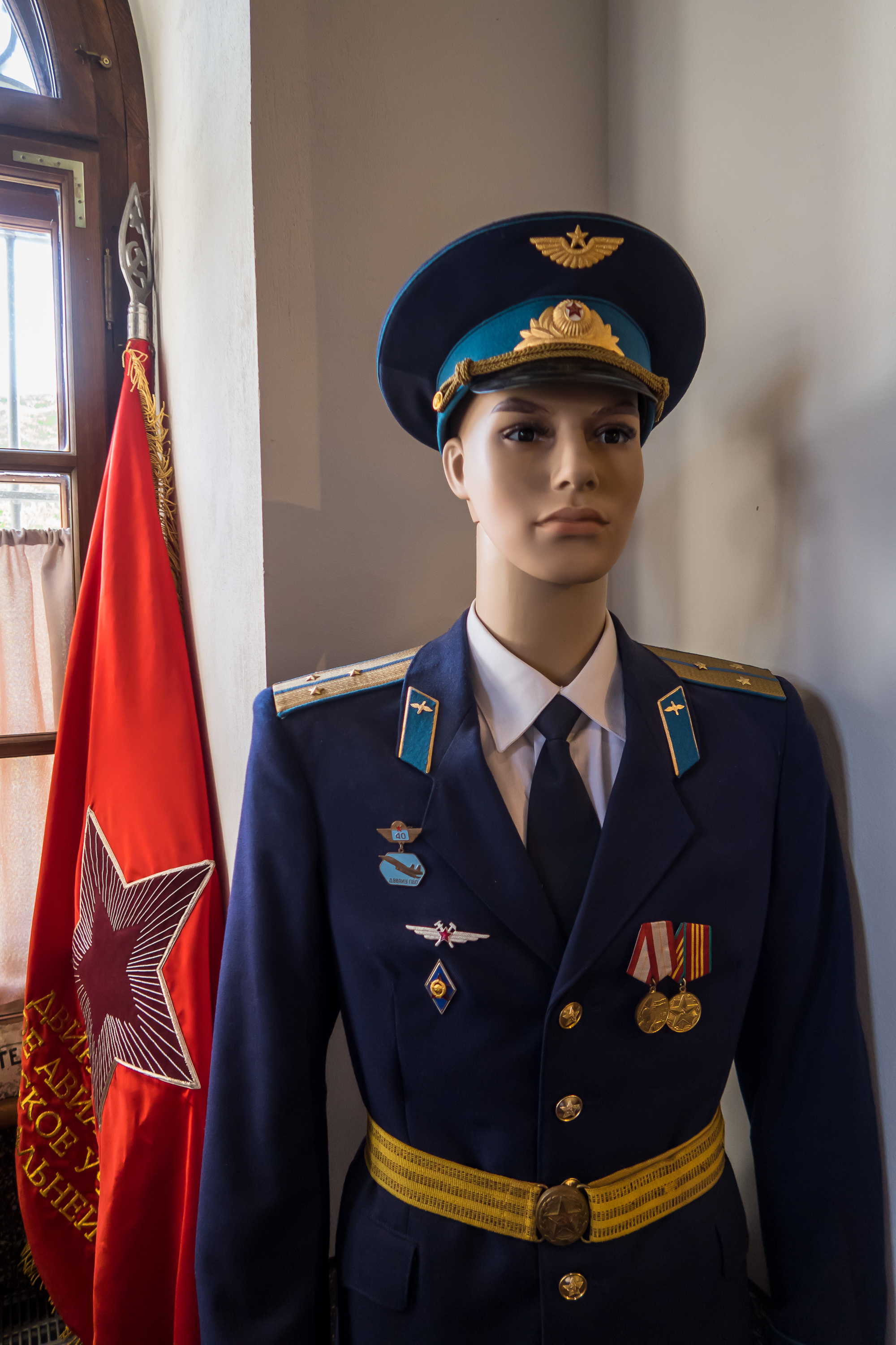 A mannequin dressed as a Soviet military officer stands indoors at the Daugavpils Fortress. The uniform is dark blue with gold accents, adorned with medals and insignia. A red flag with a large star and Cyrillic text is positioned beside it. Sunlight streams through a nearby window, creating a warm ambiance.