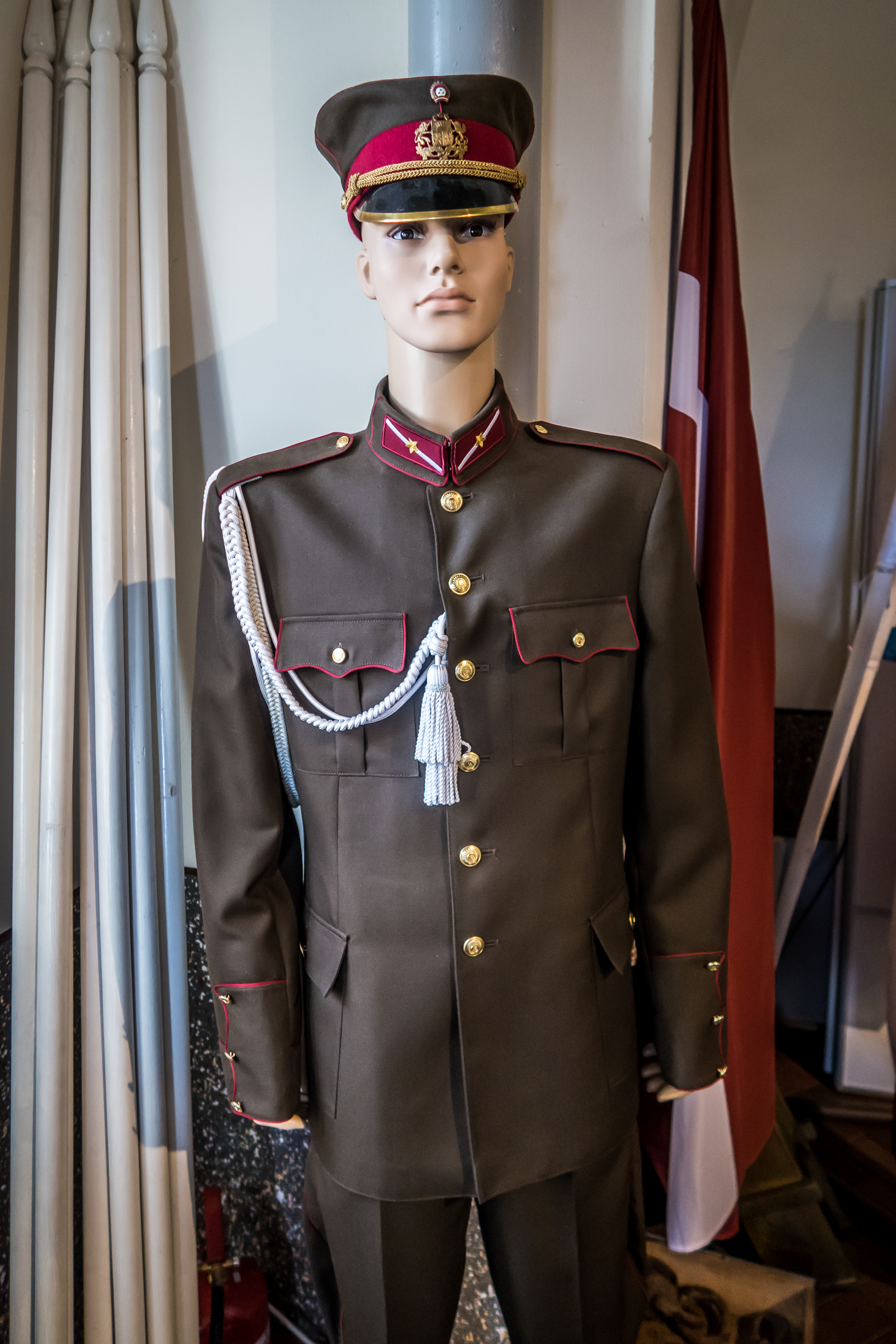 A mannequin dressed in a military uniform with a brown jacket featuring red piping, gold buttons, and an epaulet with a white aiguillette stands proudly in front of the partially visible Latvian flag. Behind it lies the historic Daugavpils Fortress, its presence enhancing this evocative display.