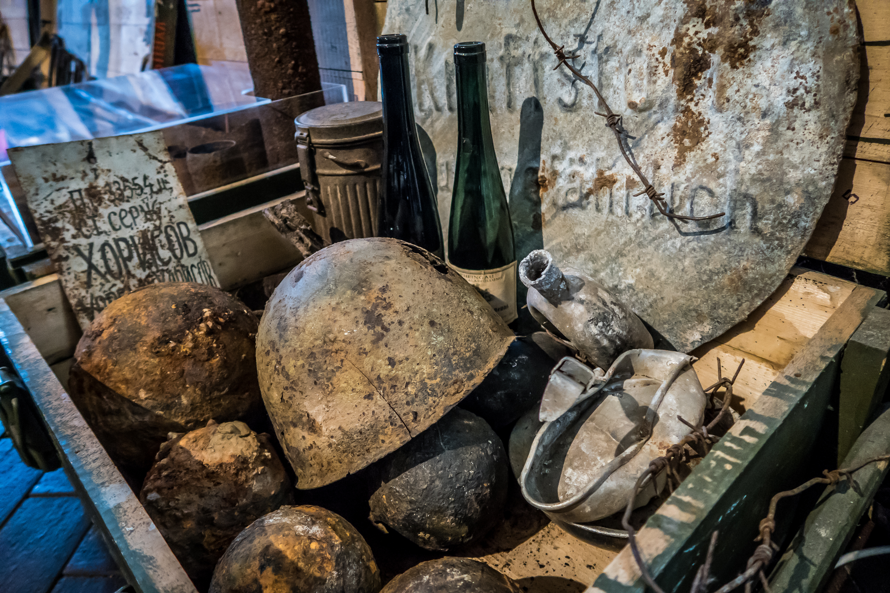 A collection of old, rusted wartime artifacts, including helmets, balls, and a circular metal sign with faded text, hints at the history of Daugavpils Fortress. Two empty green bottles and a damaged metal pitcher add to the military atmosphere in a weathered wooden crate.