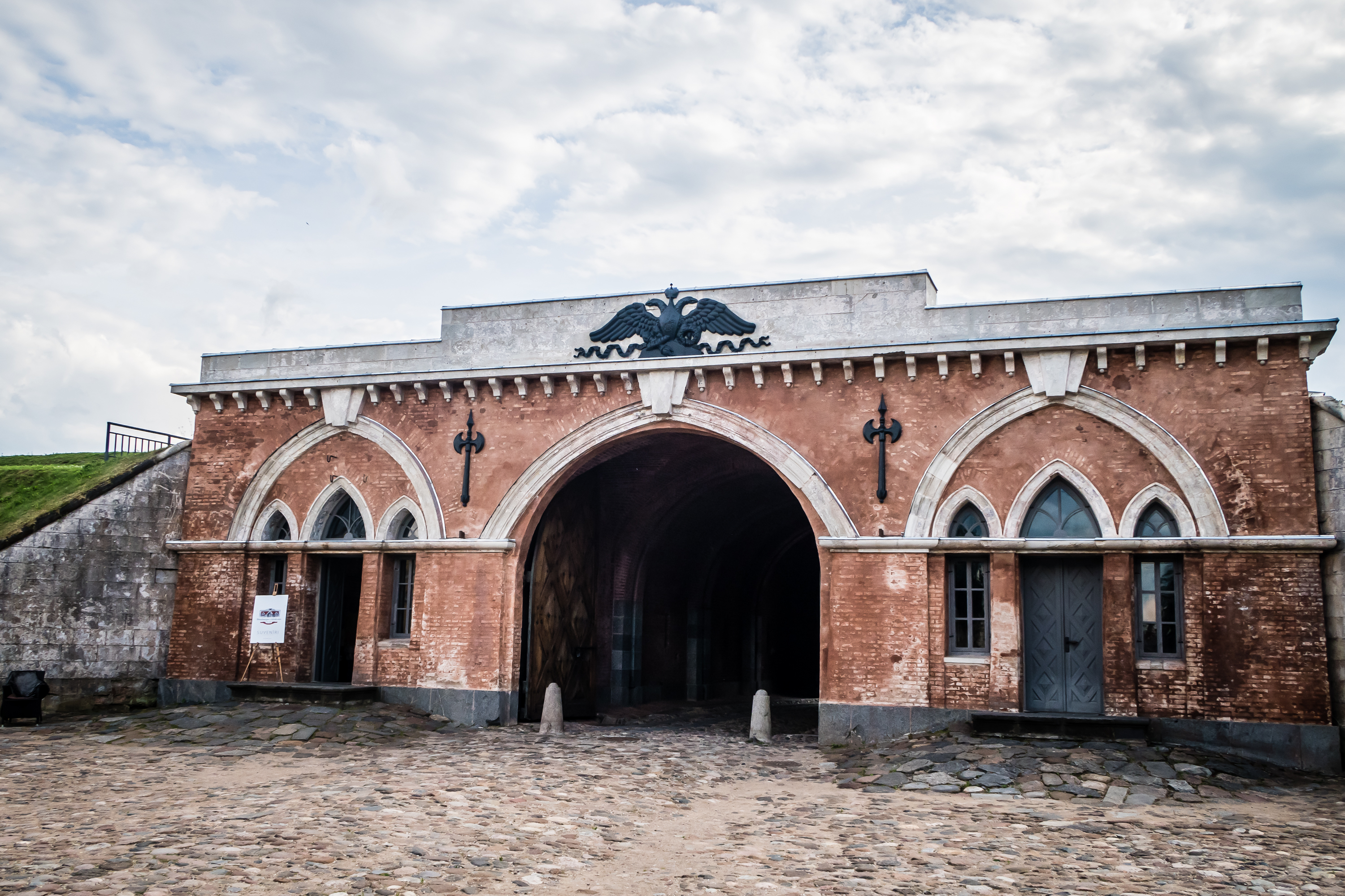 The image showcases the entrance of the historic Daugavpils fortress, featuring three arches. The central arch is taller, adorned with an ornate emblem of an eagle. Cobblestones pave the ground beneath a cloudy sky, while the two smaller side arches boast decorative trim and doors.