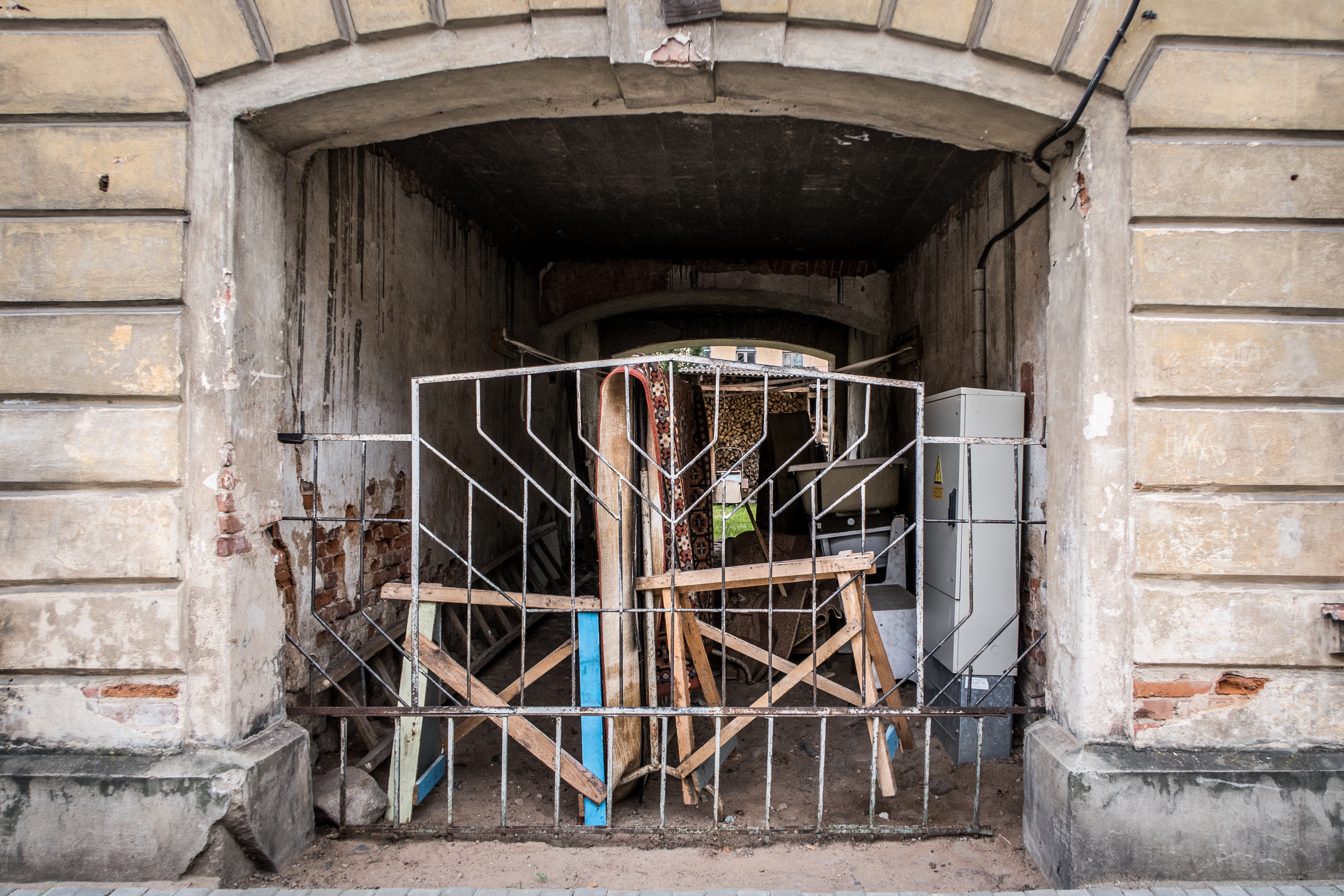 A dilapidated archway reminiscent of Daugavpils Fortress leads into a cluttered space blocked by a makeshift barrier of wooden planks and metal fencing. The surrounding walls are worn and weathered, with peeled paint and exposed brick. Inside are various items, including a refrigerator and construction materials.