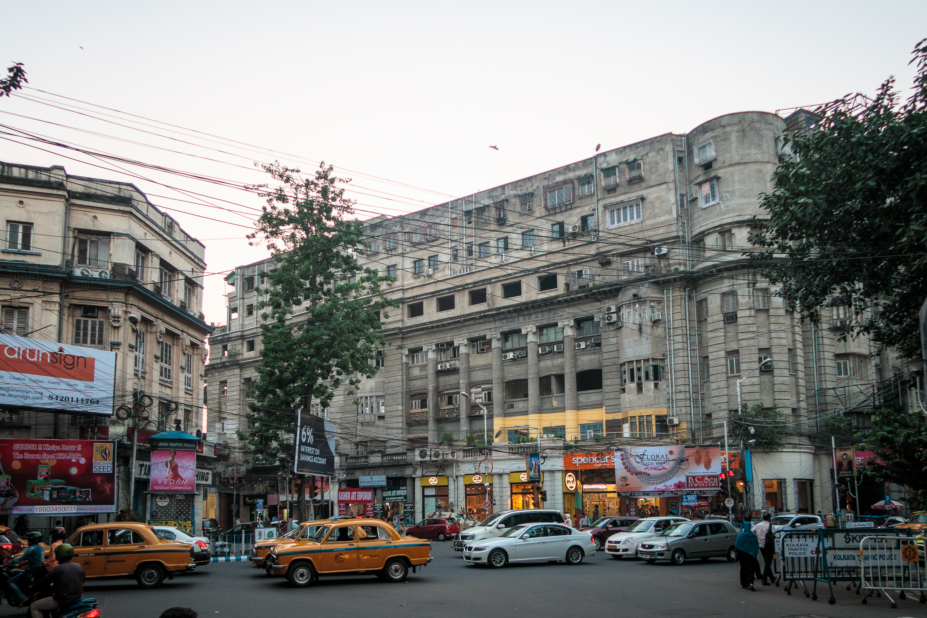 A bustling city street scene with multiple yellow taxis, cars, and pedestrians evokes the vibrant chaos of India. The backdrop features a large, grey, historic building with numerous windows and an array of colorful shop signs at ground level. The sky is clear with daylight fading; trees and power lines are visible.