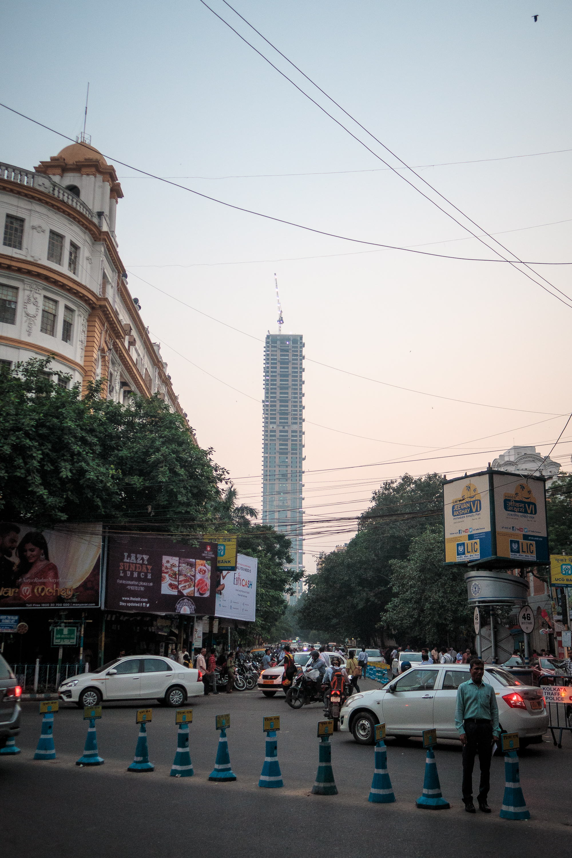 A bustling urban street intersection in India teems with people and vehicles, including cars and motorcycles, under a clear sky. A tall, modern skyscraper under construction is visible in the background, while a historic building with an ornate façade stands on the left. The street is lined with trees and various signs, including blue barriers and billboards.