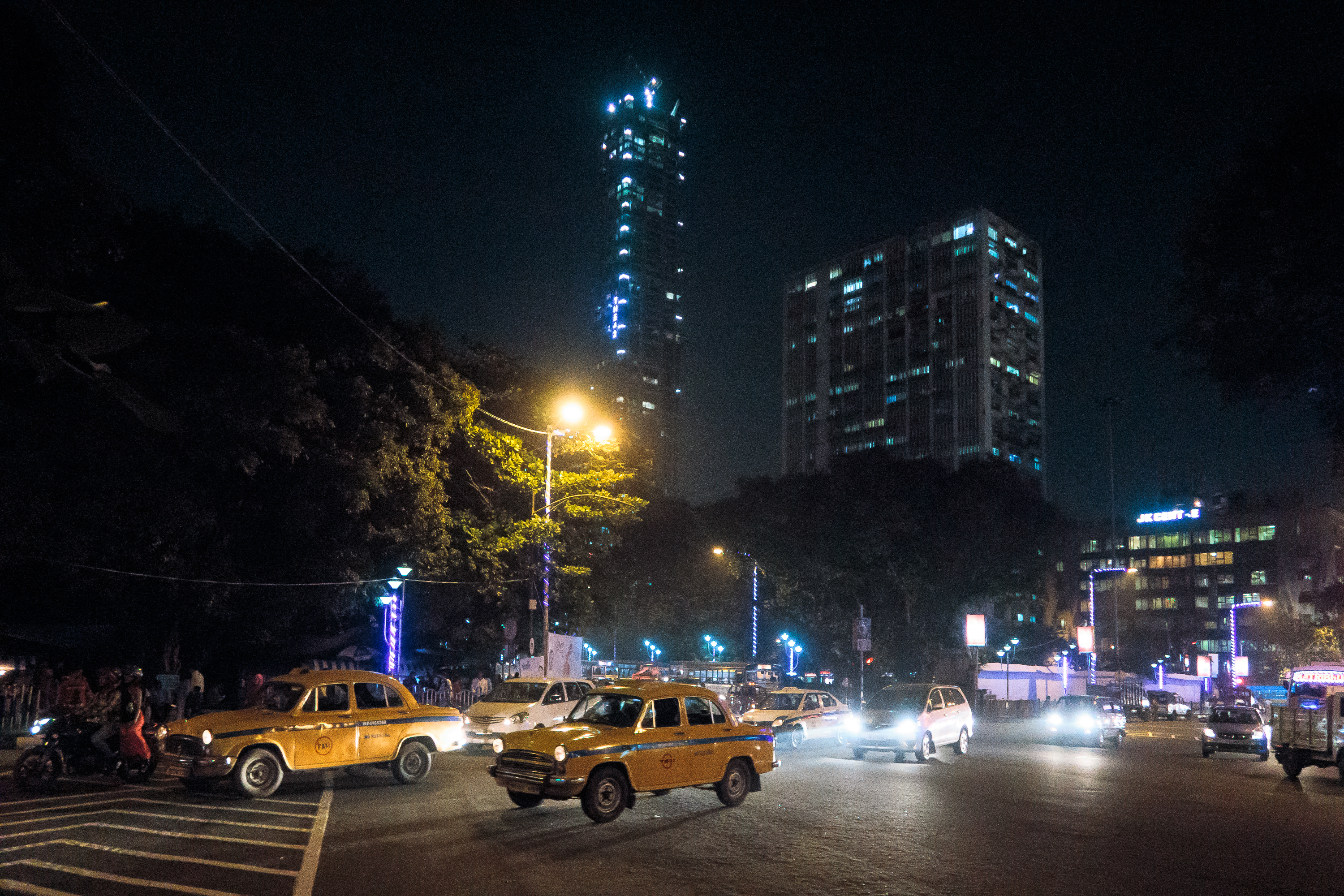 A nighttime city scene with multiple yellow taxis and cars at an intersection. Streetlights illuminate the area, and tall buildings with bright lights, including one with a distinctive blue vertical light pattern, are visible in the background. Trees line the streets, adding some greenery reminiscent of bustling cities in India.