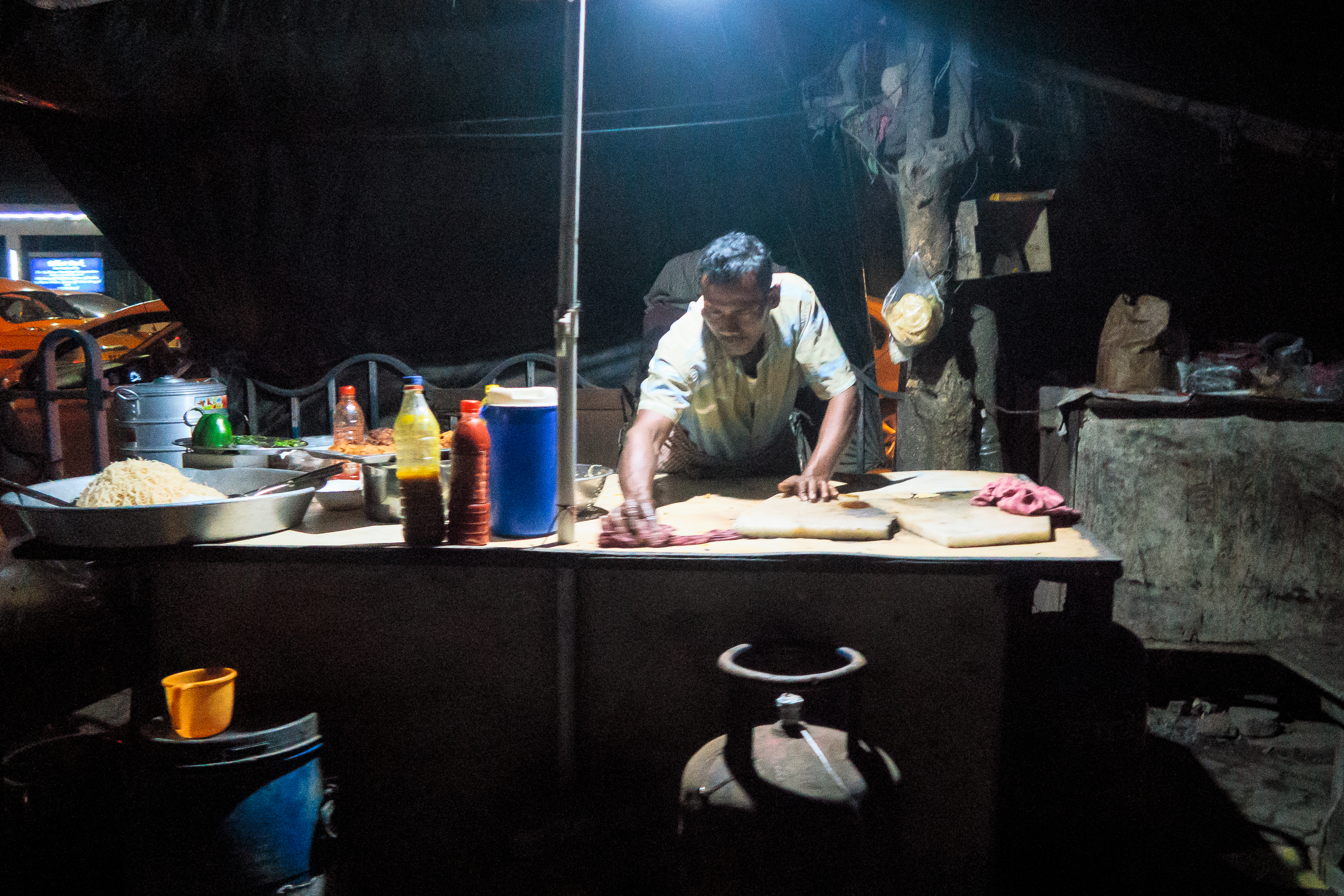 A person under a dim light works at an outdoor food stall in India. The stall features a table with several ingredients, including a bowl of noodles, condiments, utensils, and a gas cylinder beneath. A dark backdrop with hanging bags and tools forms the background, creating a rustic ambiance.