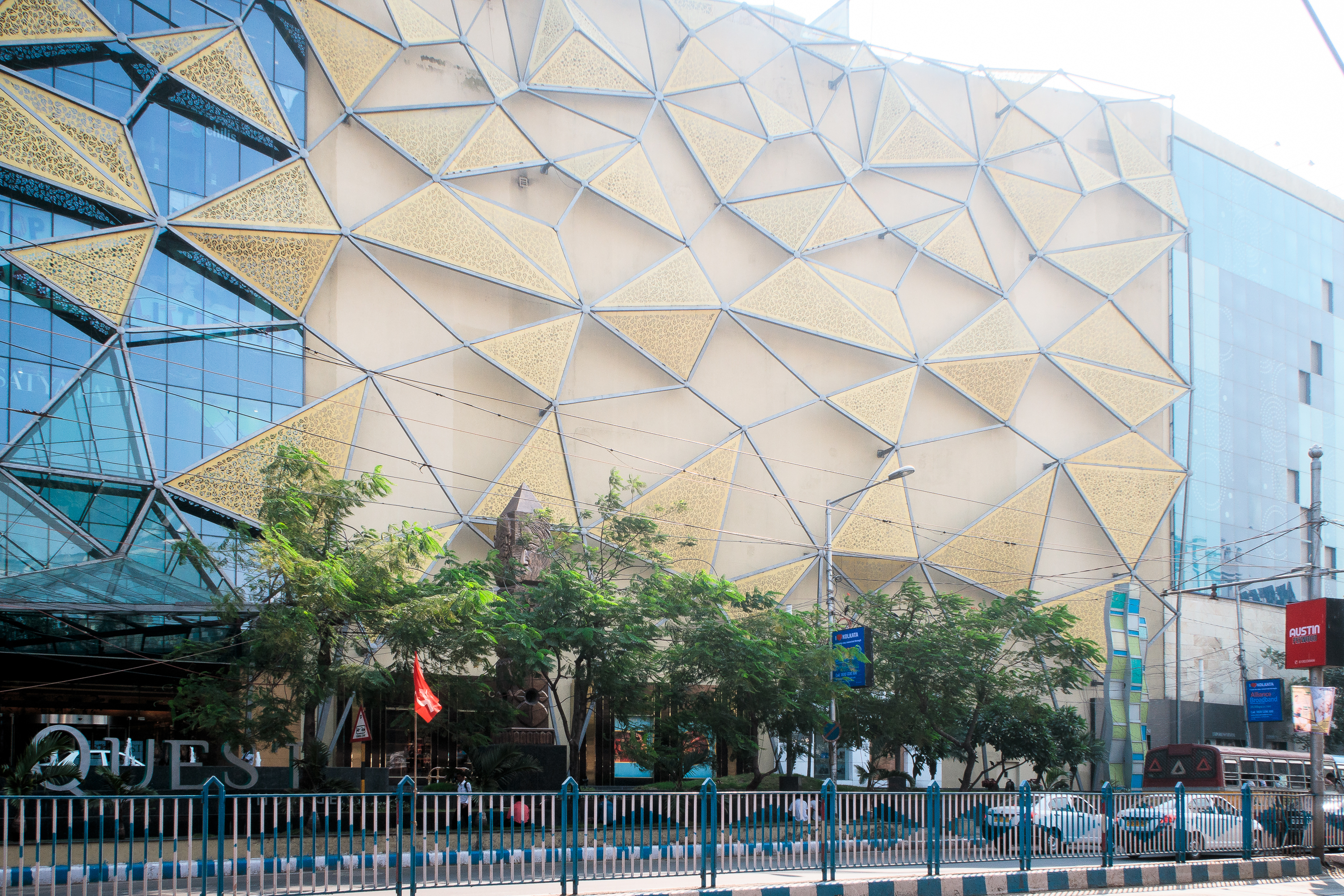 The modern building, reminiscent of architectural wonders found in India, showcases an intricate geometric facade with triangular gold and glass panels. Trees line the street in front, while traffic barriers run parallel to the sidewalk. A clear sky stretches above, dotted with some street signs nearby.