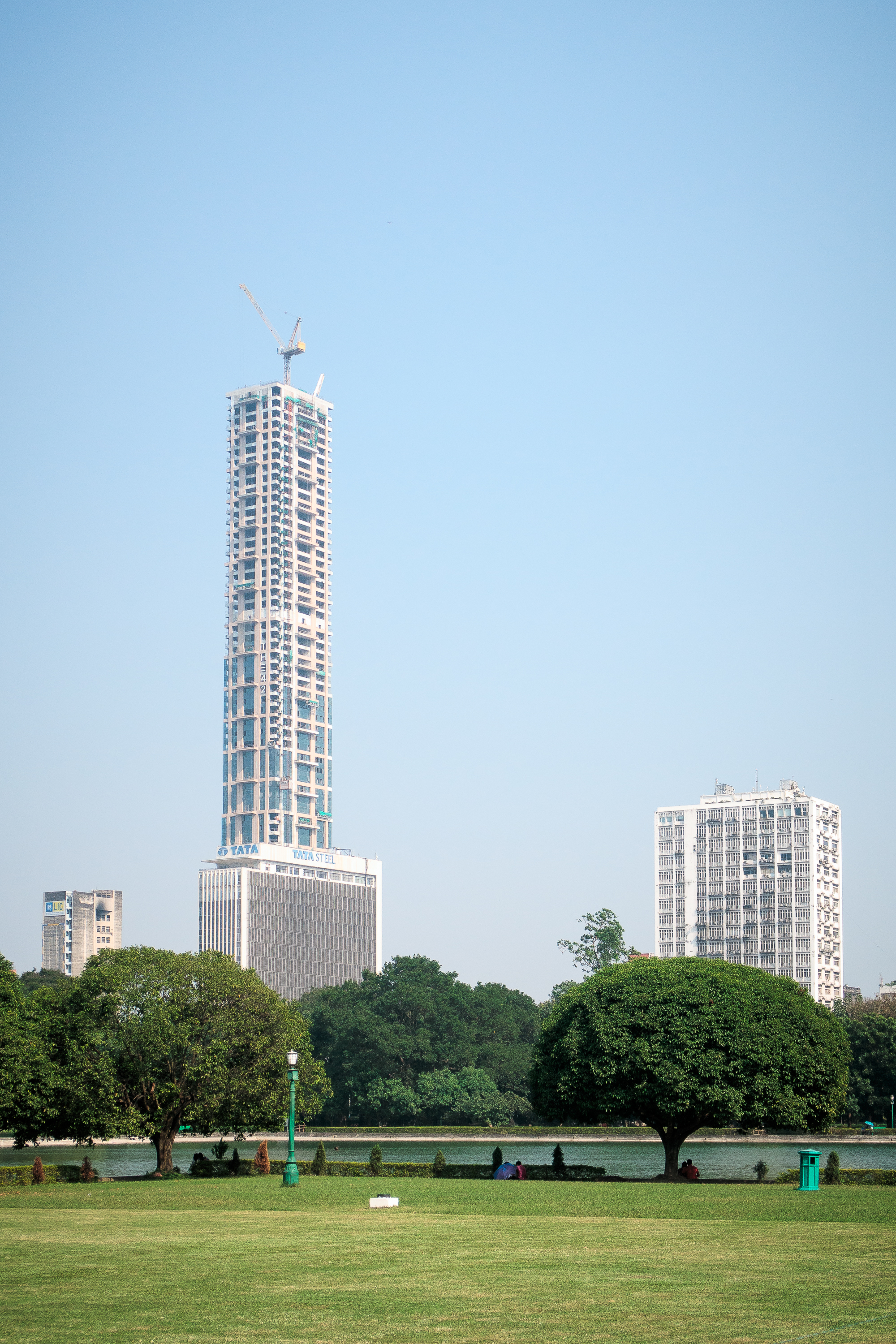 A tall skyscraper under construction with cranes on top rises majestically against a clear blue sky in India. The building is surrounded by other high-rise structures, and in the foreground, a lush green park with manicured grass hosts a few people sitting on benches.