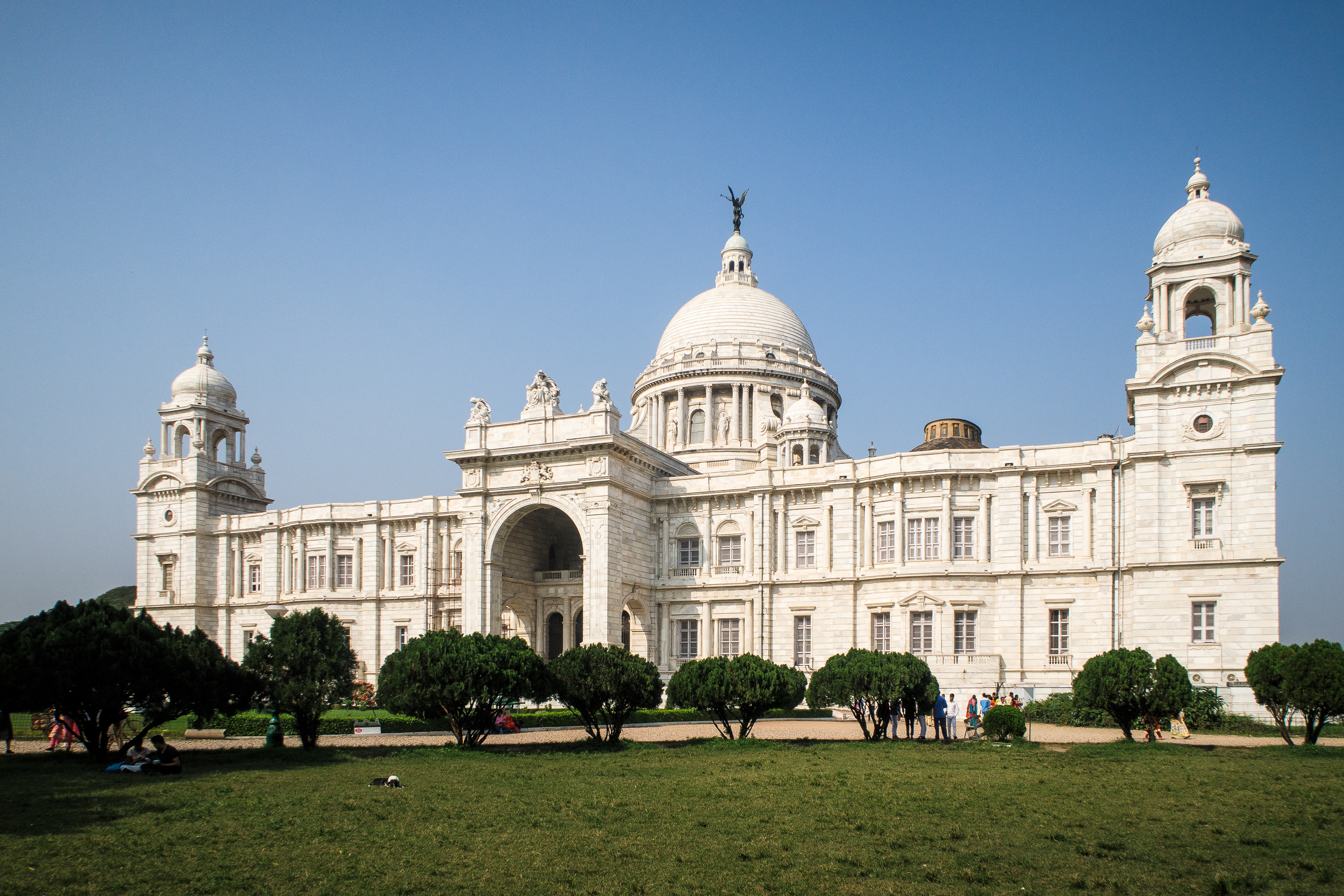 The image showcases the Victoria Memorial in Kolkata, India, a stunning white marble building with a central dome topped by a statue. Flanking it are smaller domes and towers. This iconic Indian landmark is surrounded by lush green lawns and bushes under a clear blue sky.