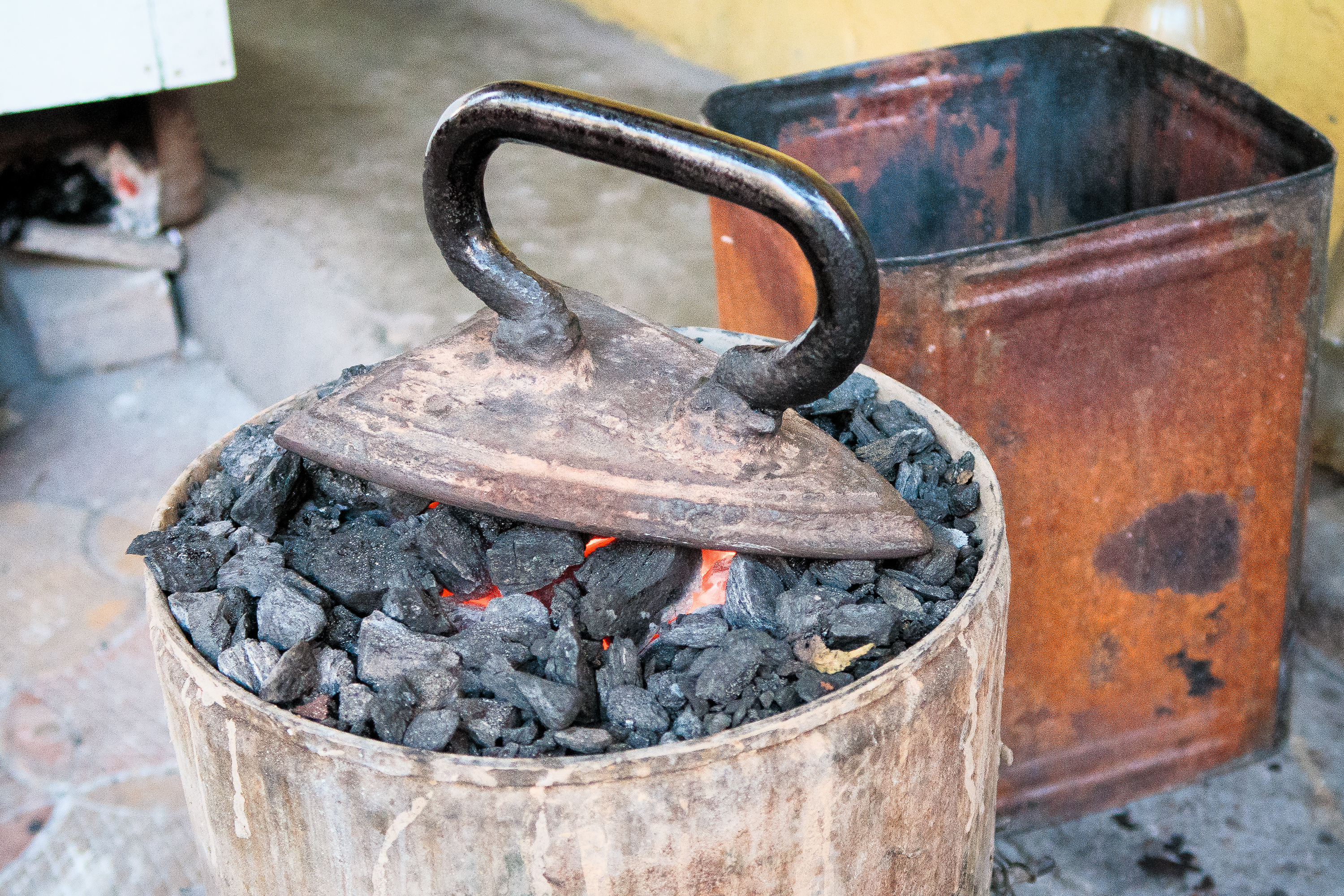 An old-fashioned coal iron rests on hot coals in a metal container, reminiscent of traditional scenes from India. Nearby, a rusted metal bin adds to the rustic setting, with concrete flooring and a yellow wall in the background. The coals glow red beneath the iron, indicating the heat used for pressing clothes.