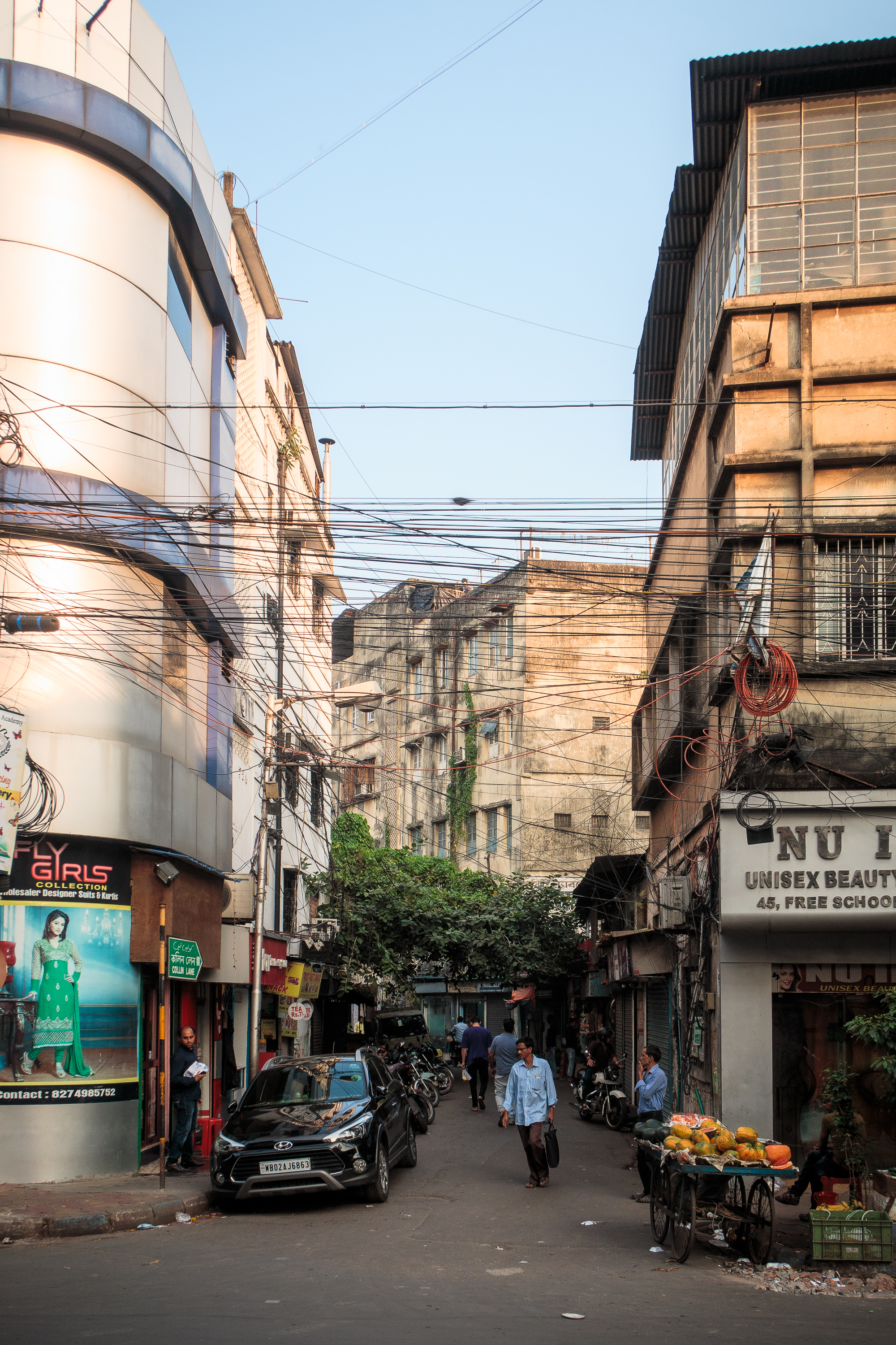 A bustling Indian urban street scene with narrow lanes, flanked by tall, weathered buildings. Shops line both sides, including a unisex beauty salon. People walk and bike through the street, and a black car is parked nearby. Overhead, many tangled electric wires cross the sky.