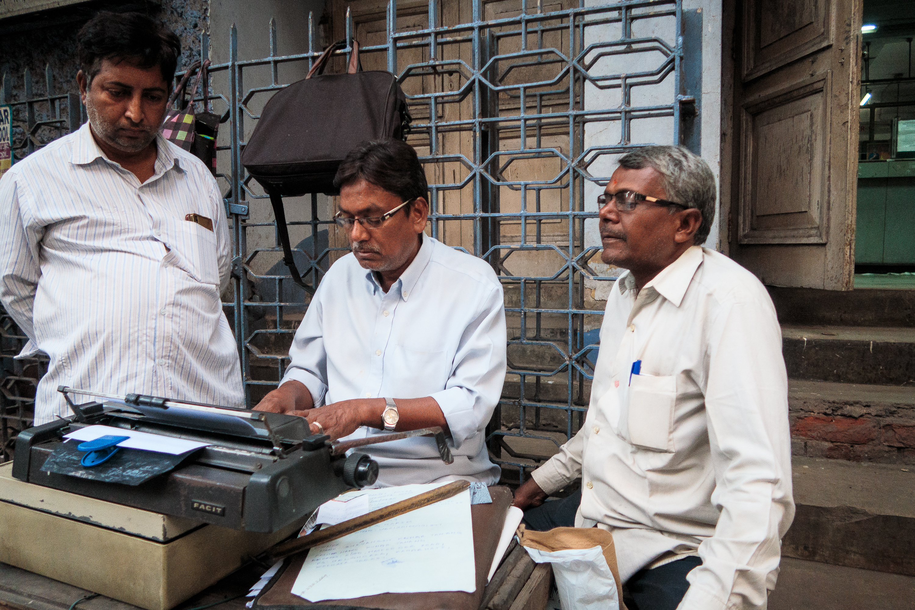 Three men are gathered around an old typewriter on a street in India. The man in the middle is typing, while the others look on. Papers and a ruler are placed on the table. They stand before a metal gate and building entrance, all dressed in light-colored, casual shirts.
