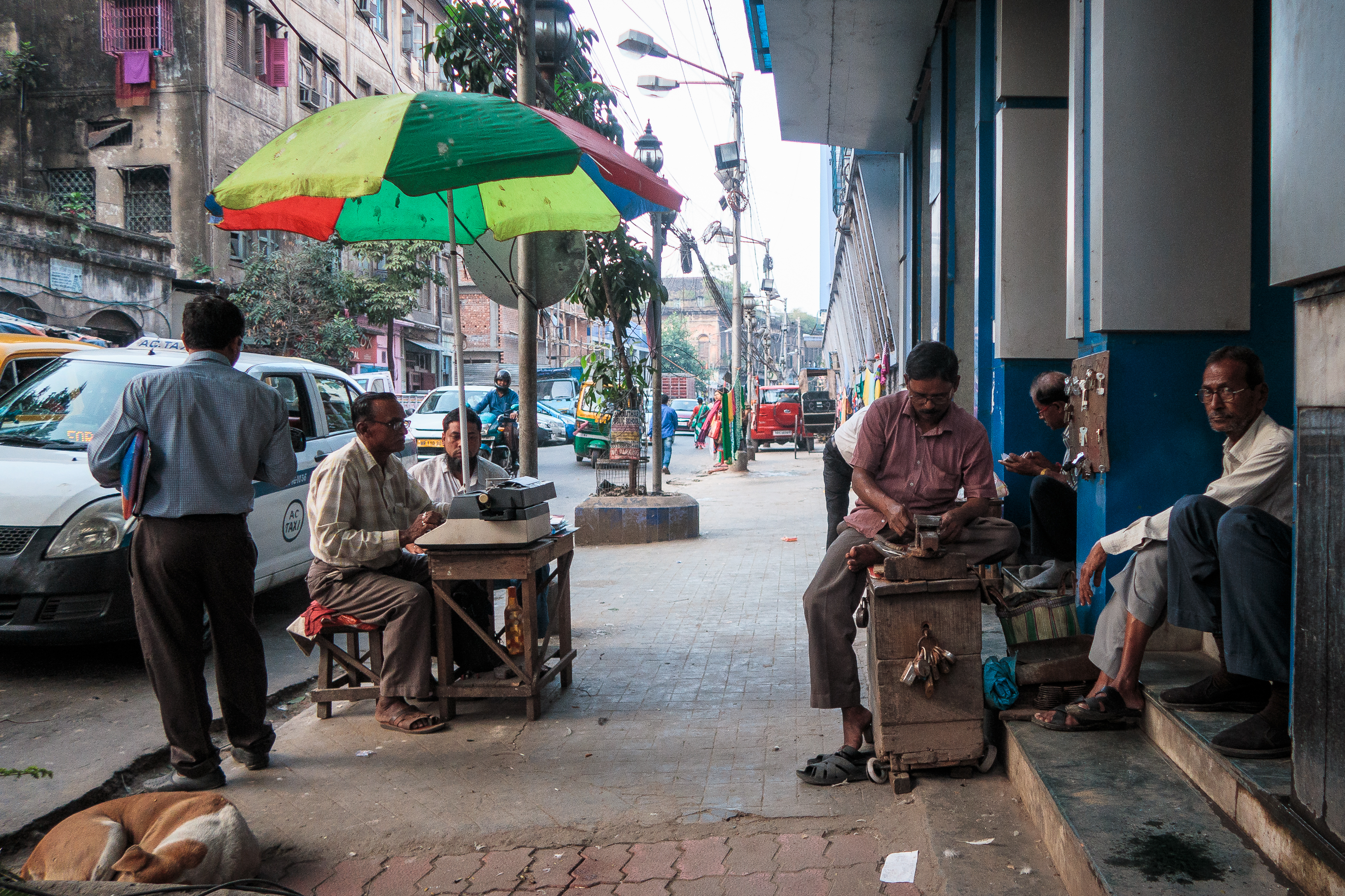 A bustling street scene in India depicts several men on a sidewalk. On the left, a man sits at a typewriter under a colorful umbrella. To the right, two men sit on steps, one with a phone. A dog lies nearby. Vehicles and buildings line the busy street, hinting at a lively urban setting.