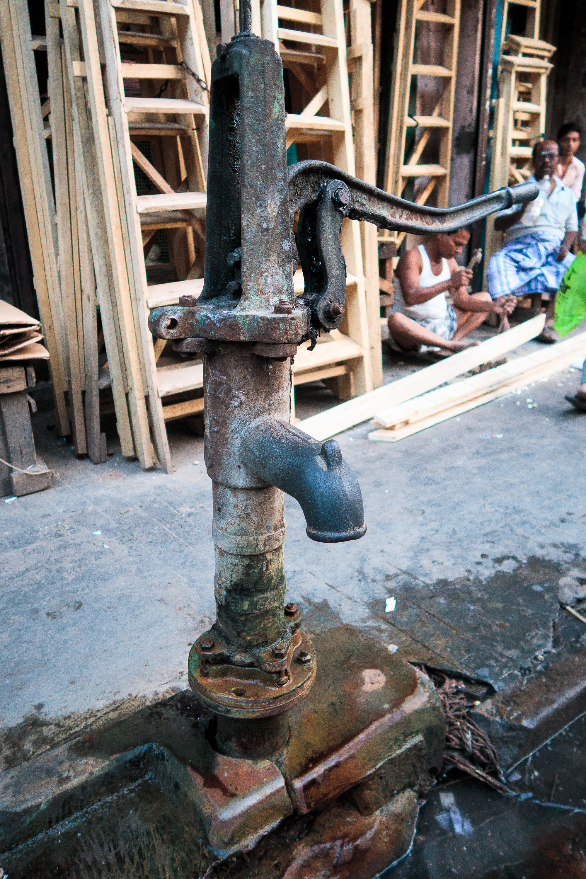 A rusty, old-fashioned hand water pump stands on a stone base in an Indian street. Behind it, several wooden frames are stacked vertically. In the background, a group of men sit on the ground, relaxing and chatting, with one wearing a traditional white dhoti. The setting is urban and informal.
