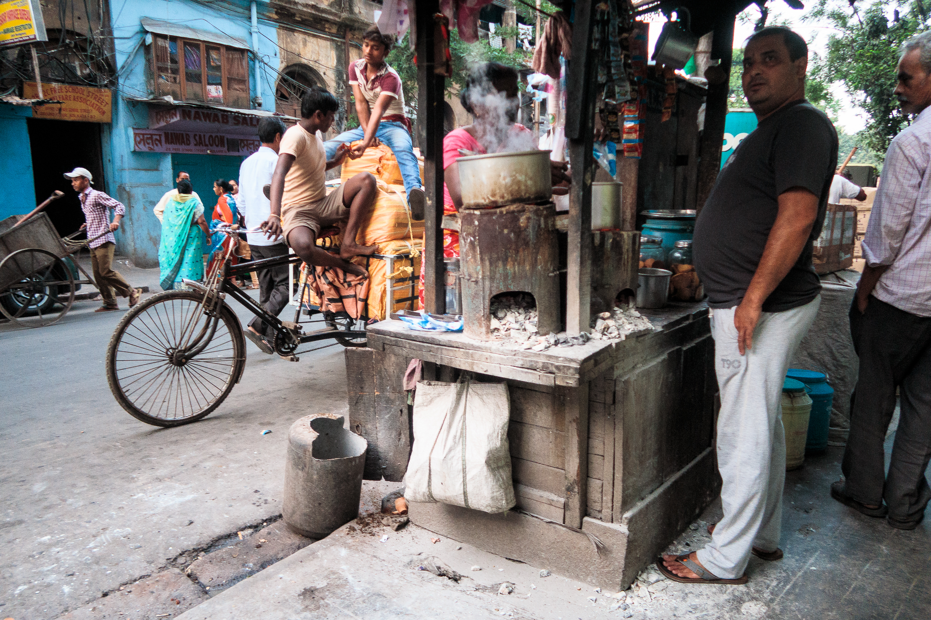 A bustling street scene in a vibrant Indian market area. A roadside vendor stands by a traditional clay stove with pots steaming. A bicyclist rides by, and a man sits on a rickshaw. The backdrop features colorful buildings and pedestrians walking past in various attire.