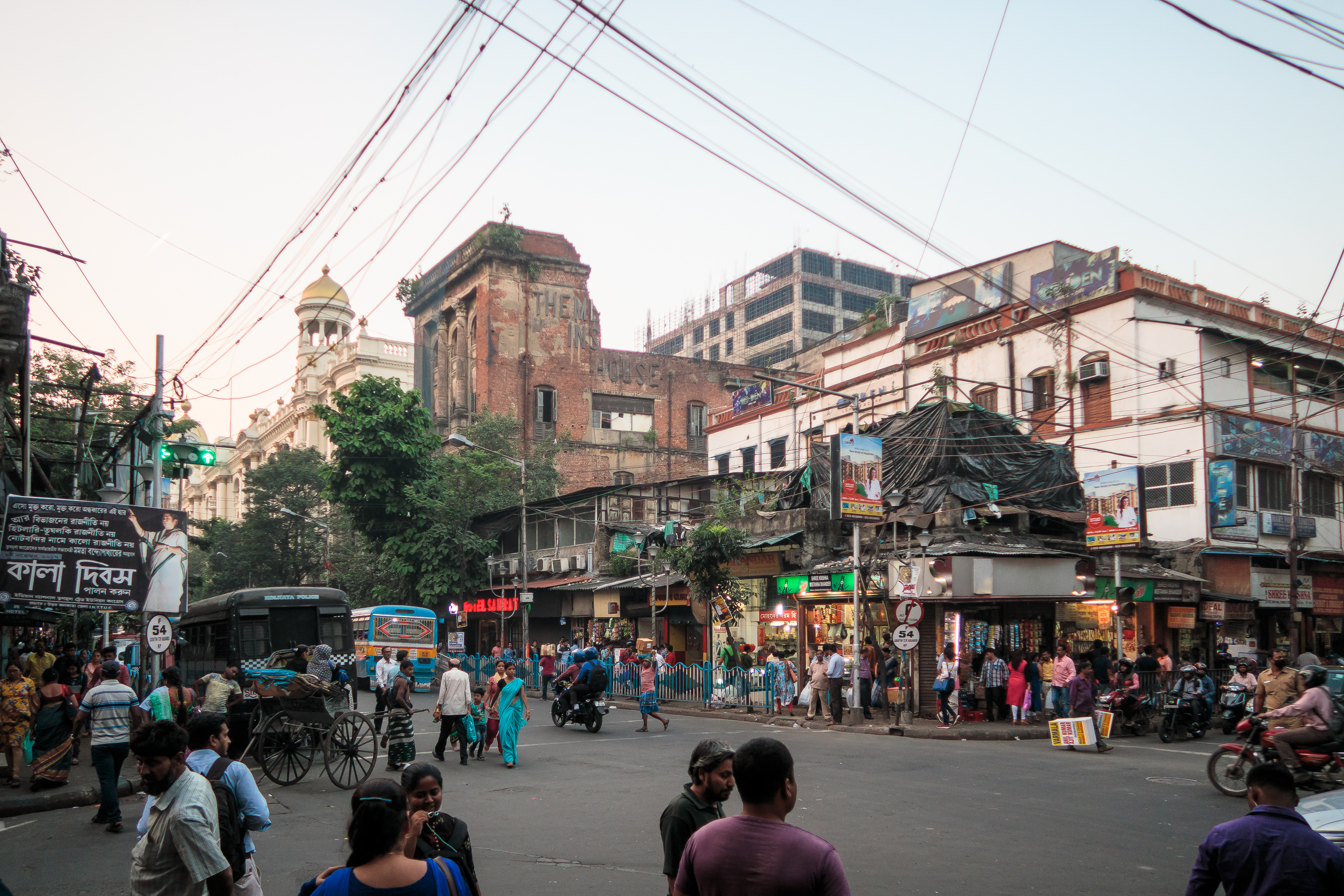 A busy street scene in Kolkata, India, with numerous pedestrians, rickshaws, and vehicles. The vibrant backdrop showcases a mix of historic and modern Indian buildings, including a large brick structure. Overhead wires crisscross the scene amidst various shop signs and billboards.