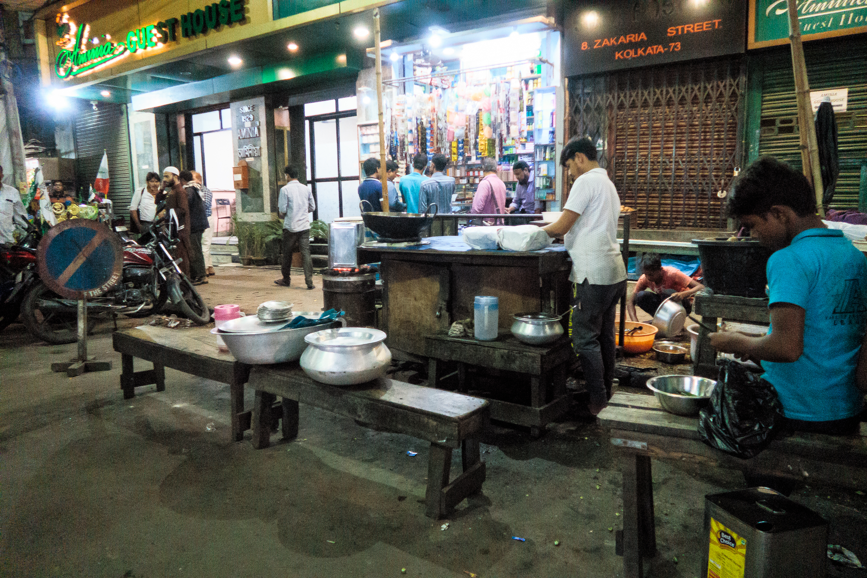 Street scene in India showing people in front of a restaurant. Two men wash dishes under dim lights while motorcycles are parked nearby. A shop with colorful items is visible in the background, and the street sign reads "ZAKARIA STREET." There’s a no-entry traffic sign on the left.