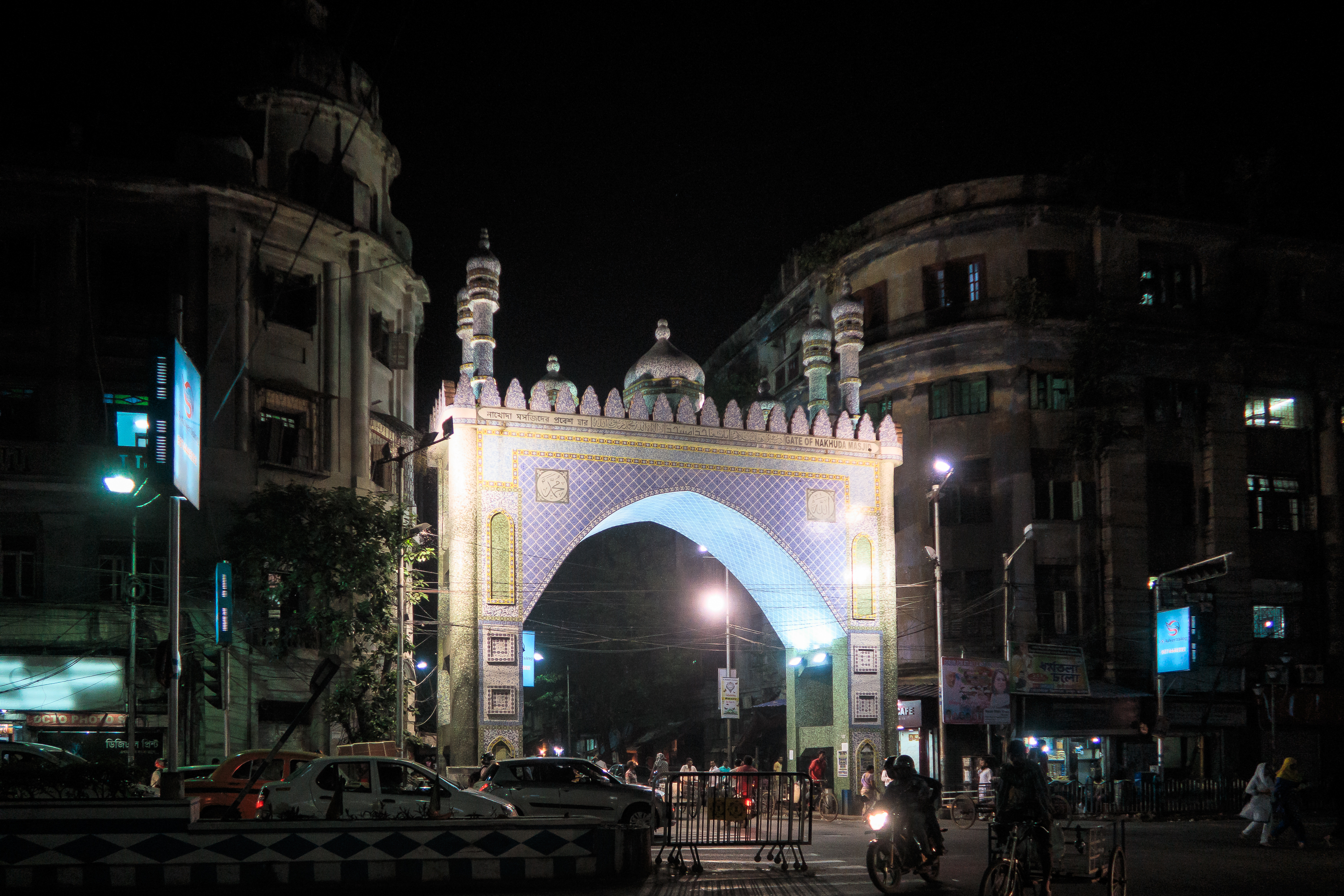 A night view of an illuminated gateway with intricate designs, reminiscent of India's architectural charm, resembling an arch with minarets. The lively scene includes people walking and cars passing by, while the dimly lit buildings around accentuate the brilliance of the bright arch.