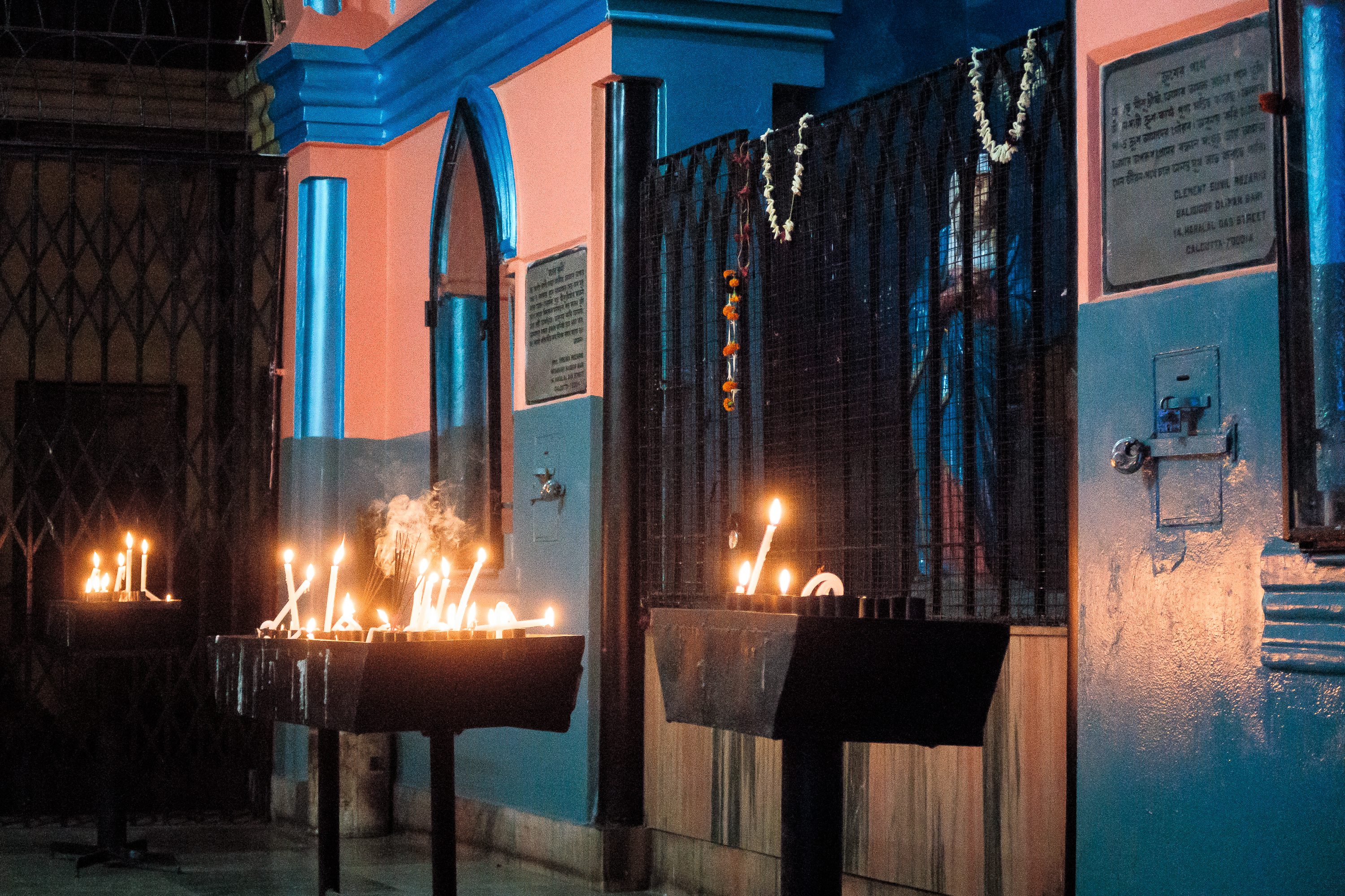 Candles are lit on stands in front of a building with blue and orange walls, reminiscent of vibrant India. The entrance features arched windows, and garlands hang on black metal bars. Plaques or signs adorn the wall, while the dim lighting creates a warm and peaceful ambiance.