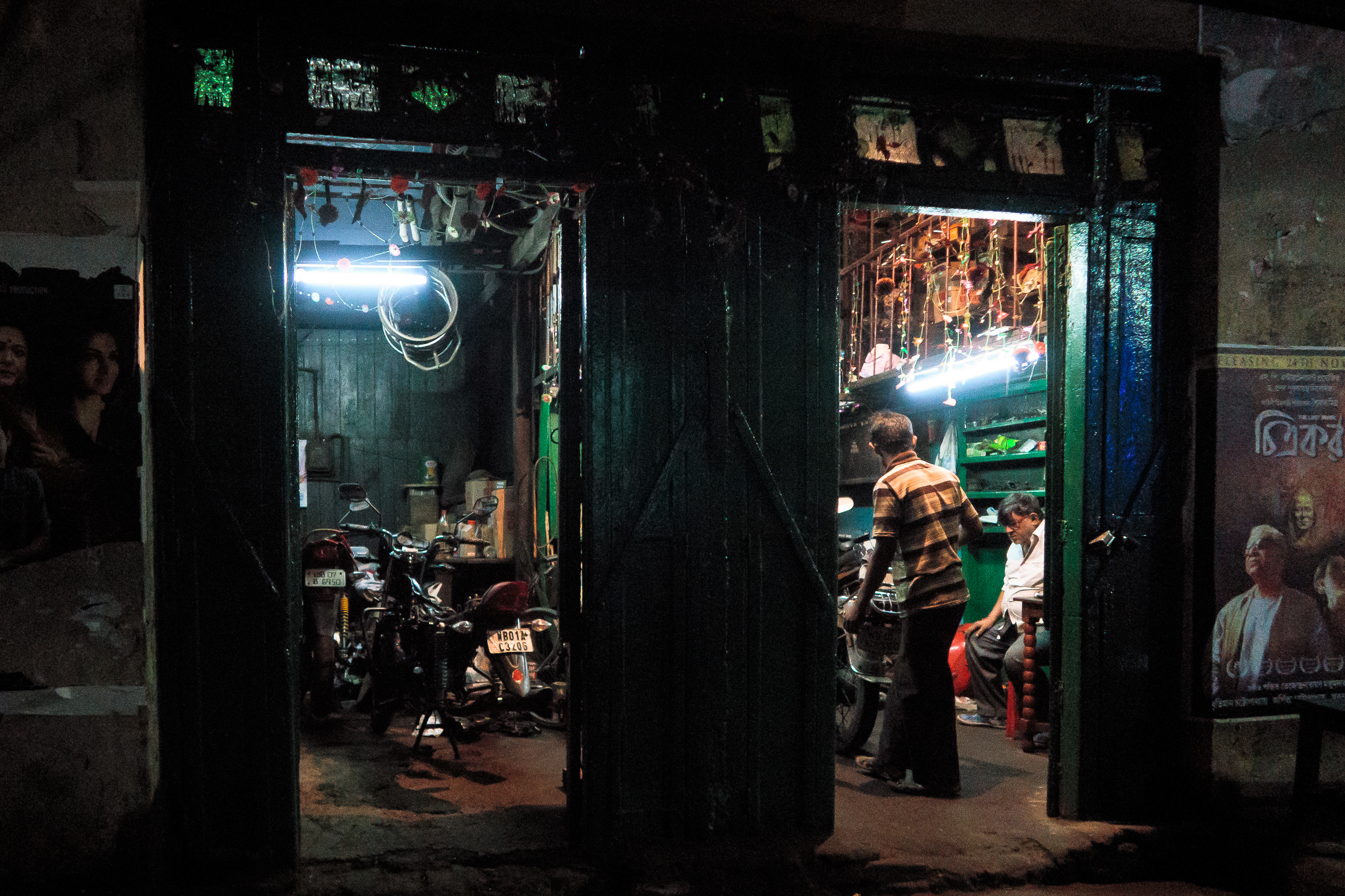 In a dimly lit garage reminiscent of India's bustling backstreets, two open doors reveal motorcycles inside. A man in a striped shirt stands nearby, while another sits inside. The walls are adorned with tools, and fluorescent lights illuminate the space, casting shadows over posters on the exterior wall.