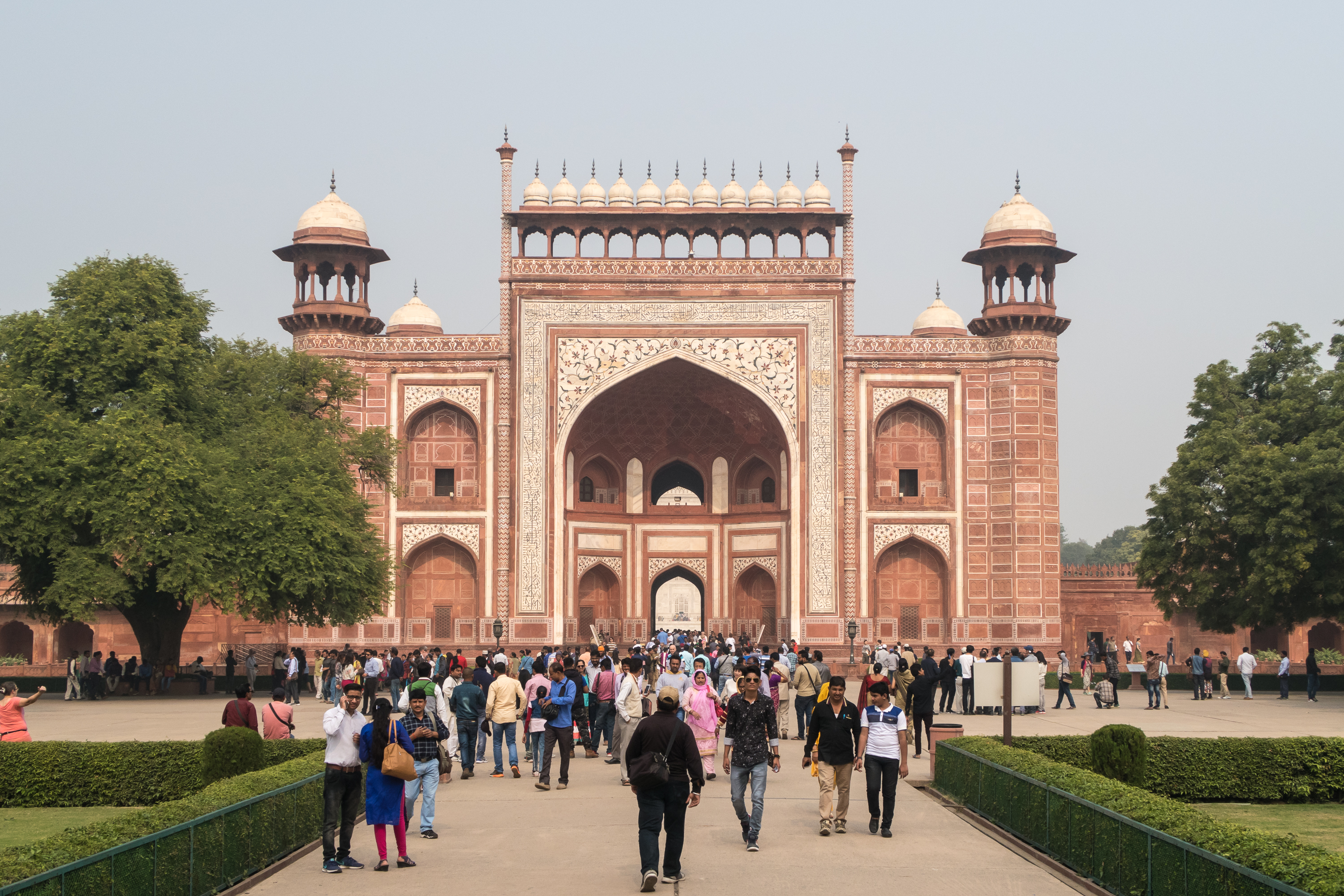 The image shows a large group of people walking towards the Great Gate (Darwaza-i-rauza) entrance of the Taj Mahal in Agra, India. The gate is an elaborate structure with intricate red and white decorative patterns, arches, and minarets. Lush green trees and manicured gardens flank the path.