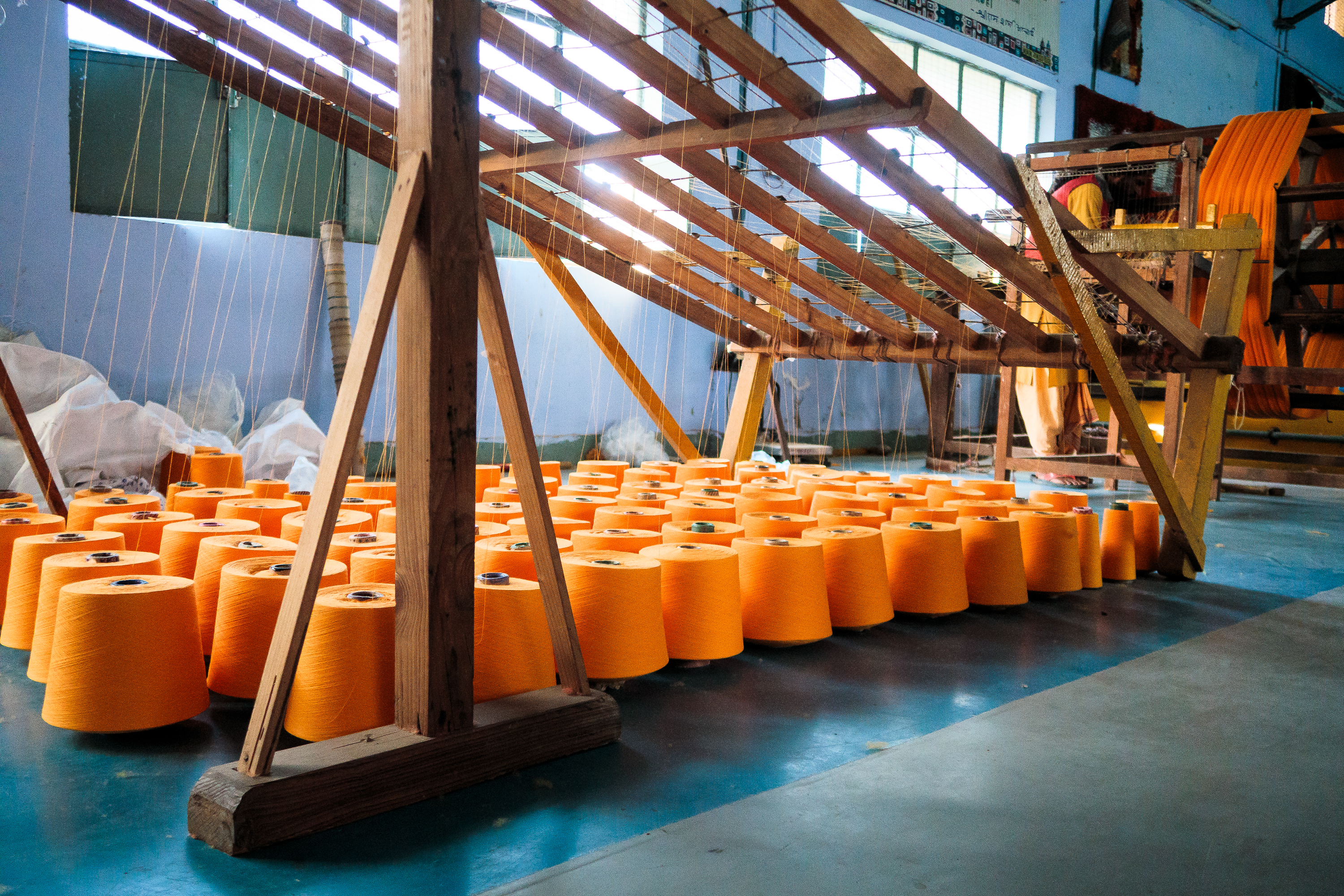 Orange spools of thread are neatly arranged in rows on a blue-painted factory floor in India. A wooden loom stands in the background, and a worker can be seen operating machinery. The well-lit setting highlights the orderly setup and equipment essential for textile production.