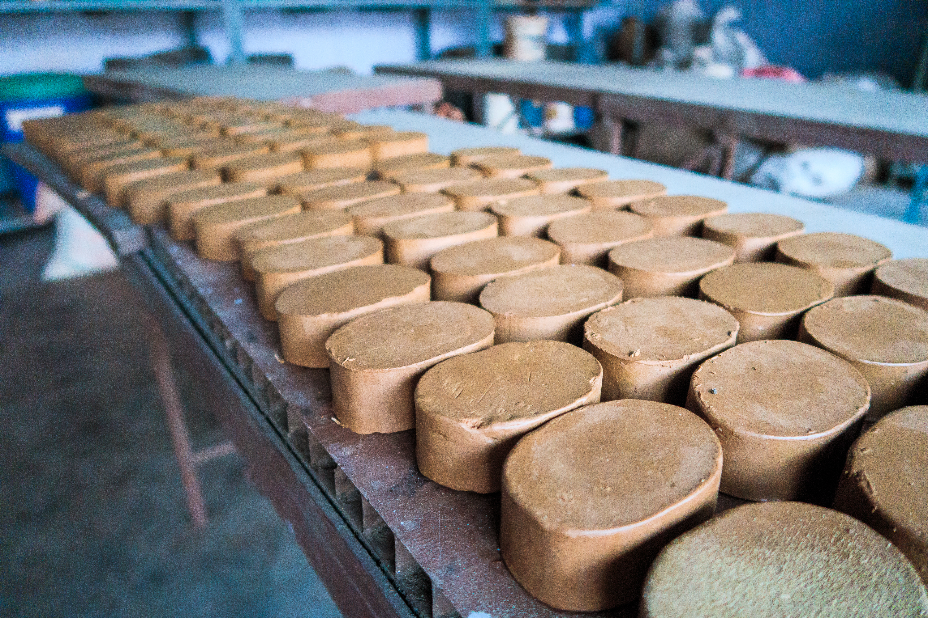 Rows of cylindrical clay pieces are neatly arranged on a wooden table in an Indian workshop setting. The smooth, brown surfaces reflect light from a window, creating a slight sheen. Shelves and various tools are blurred in the background, indicating a vibrant workspace for pottery or sculpture.