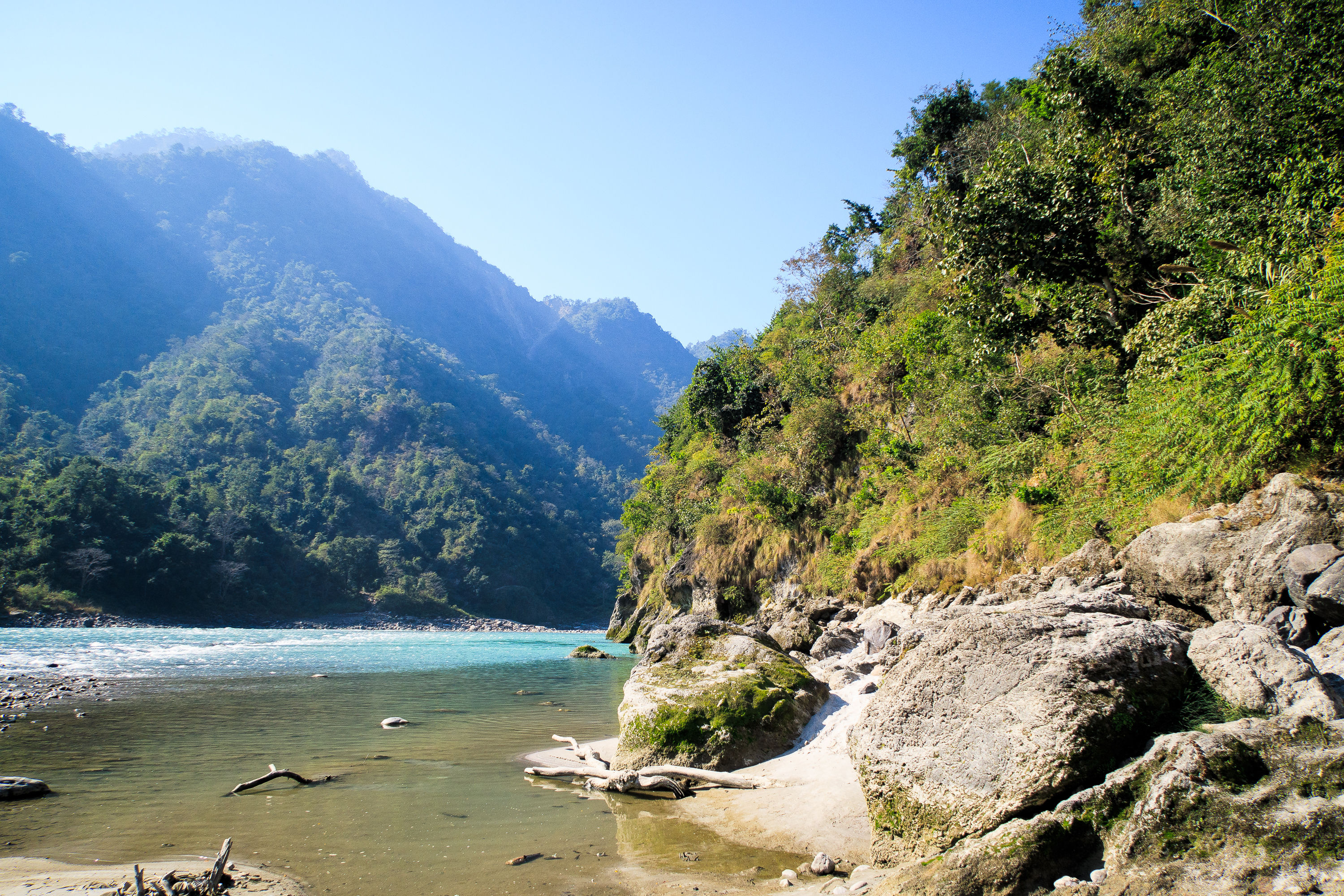 A serene riverside landscape in India with clear blue skies. The river flows gently, bordered by rocky and sandy shores. Lush green hills rise in the background under bright sunlight. The scene is tranquil, with clear water reflecting the greenery and sky.