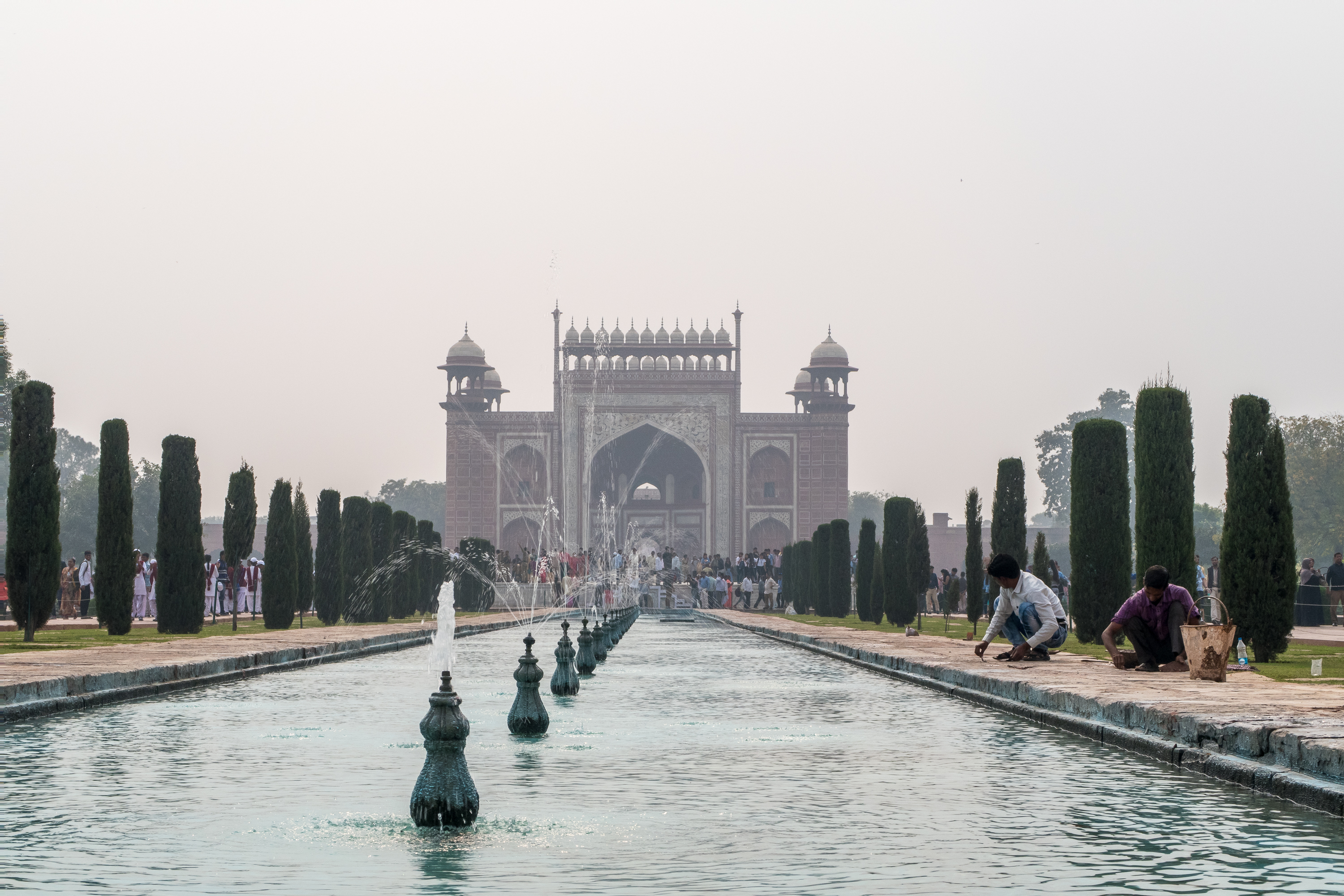 A wide-angle shot of a serene garden in India with a long rectangular reflecting pool leading to a grand Mughal-style gateway in the distance. The pool is adorned with ornate fountains, lush green cypress trees flank the pool, and people are visible near the gateway and poolside, some tending to the garden.