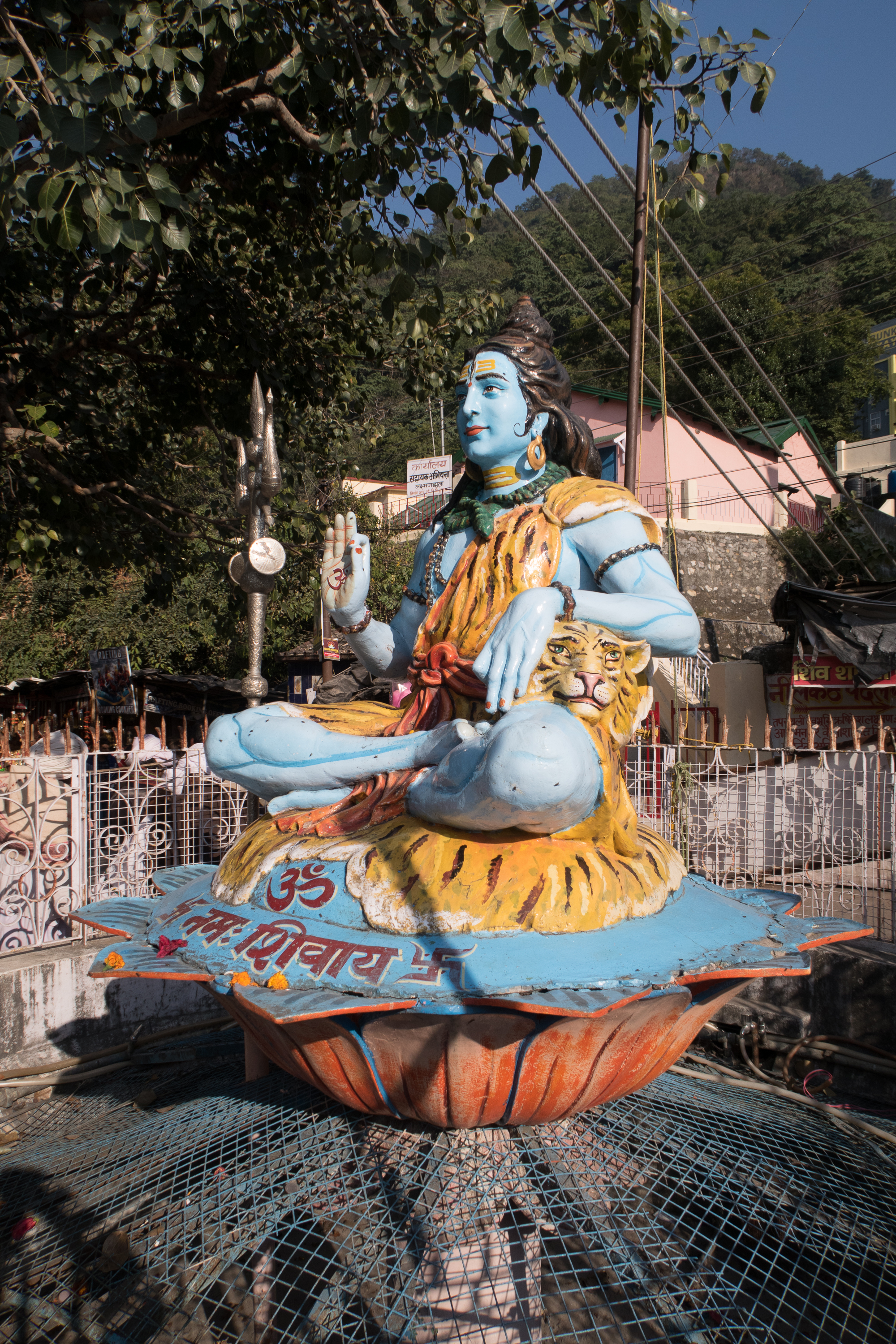 A vibrant statue of Lord Shiva in a meditative pose, painted blue and adorned with a tiger skin garment, is seated on a lotus pedestal. The deity holds a trident and has a serene expression. This iconic symbol of India is encircled by metal fencing with trees, buildings, and power lines in the backdrop.
