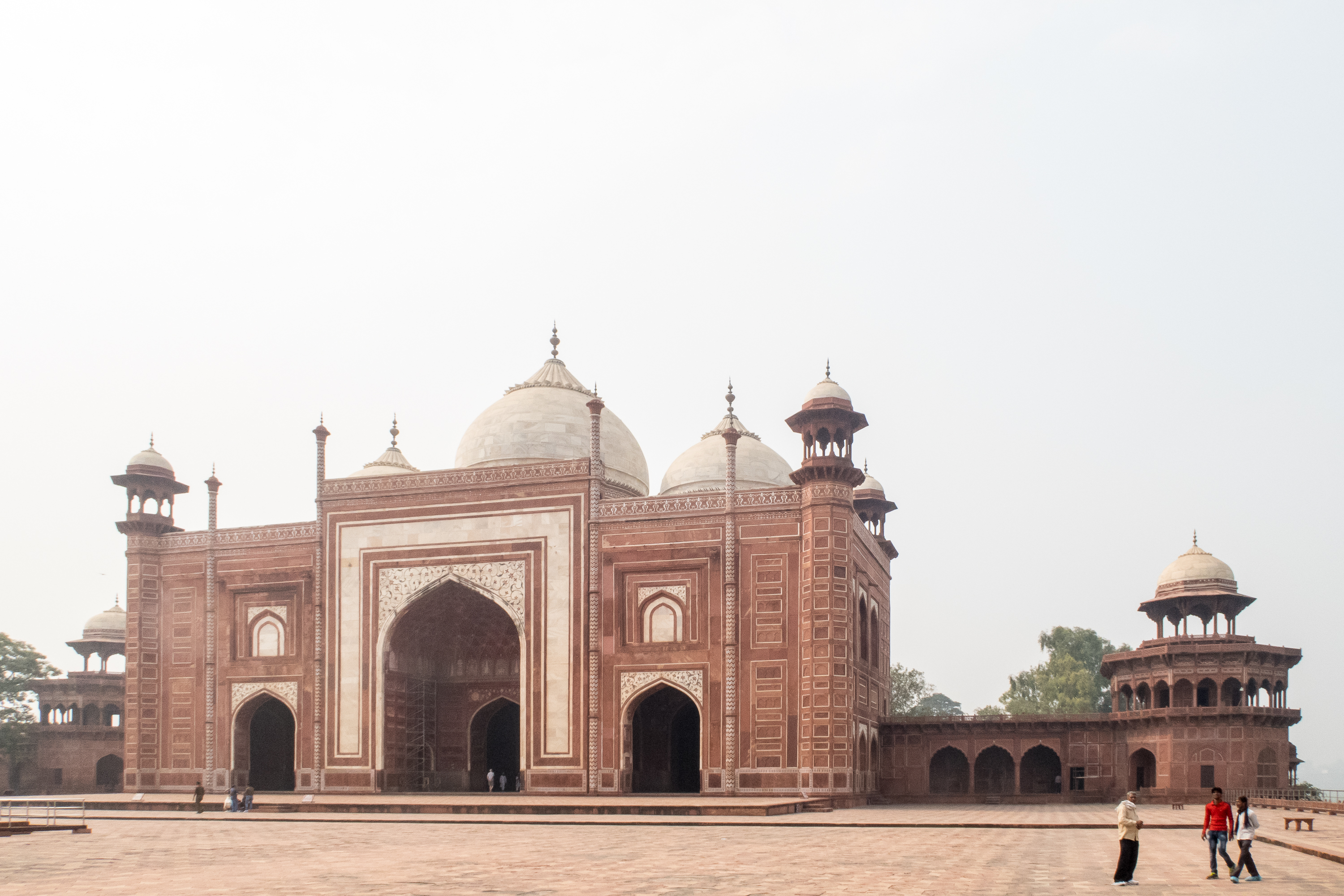 The image shows the Taj Mahal mosque in India, made of red sandstone with intricate white marble inlay. It features three domes and four minarets. Three people walk in the foreground on the expansive courtyard, two wear beige and red and one in red and blue. The sky is clear and light.